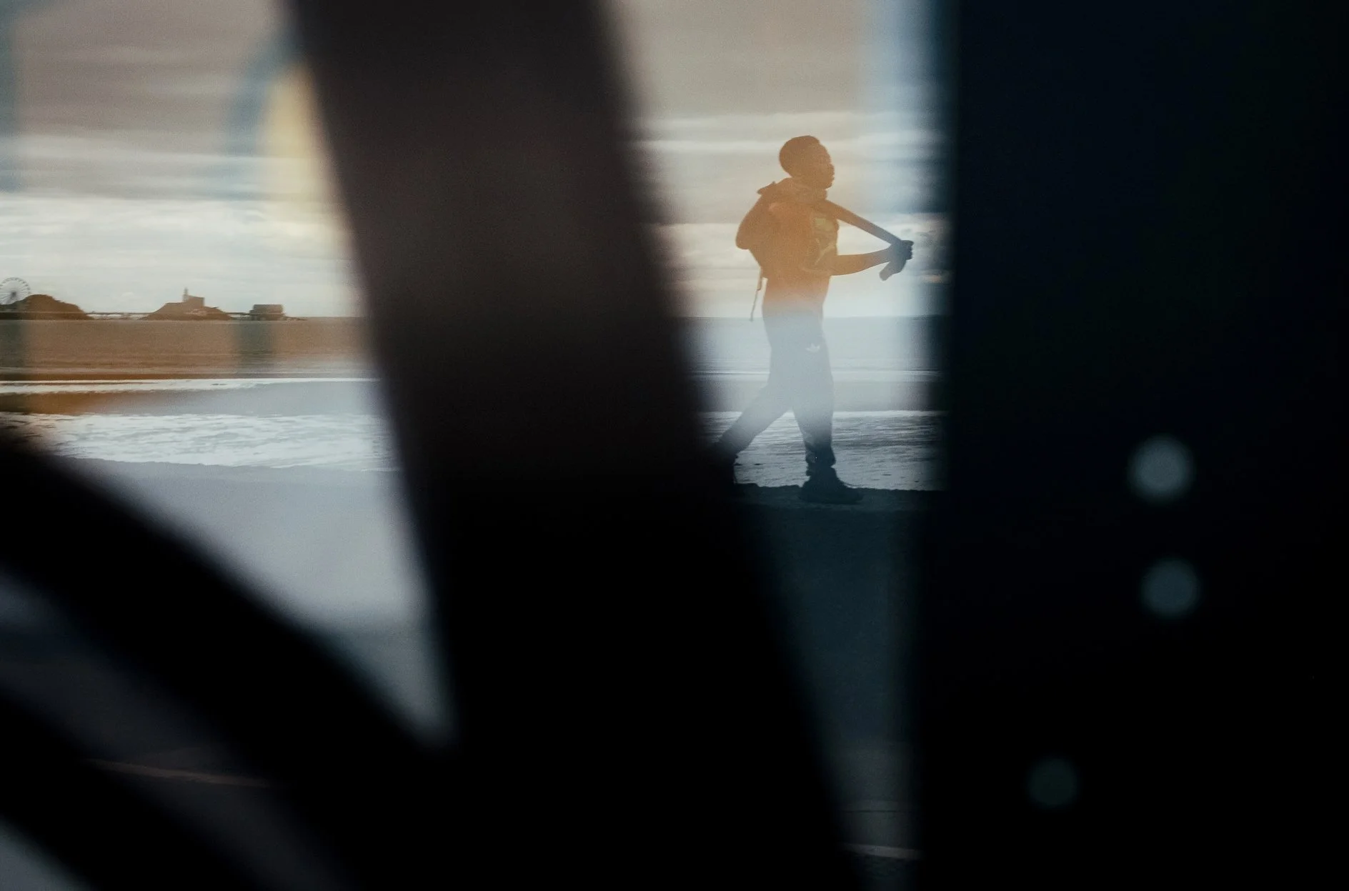 Silhouette of a person standing on the beach holding a skateboard, viewed through a window with blurred foreground objects.