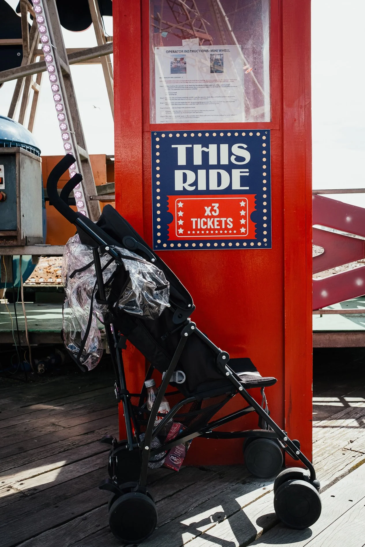 A colorful amusement park ride ticket booth with a sign that reads 'This Ride X3 Tickets' and a stroller parked next to it on a wooden boardwalk.