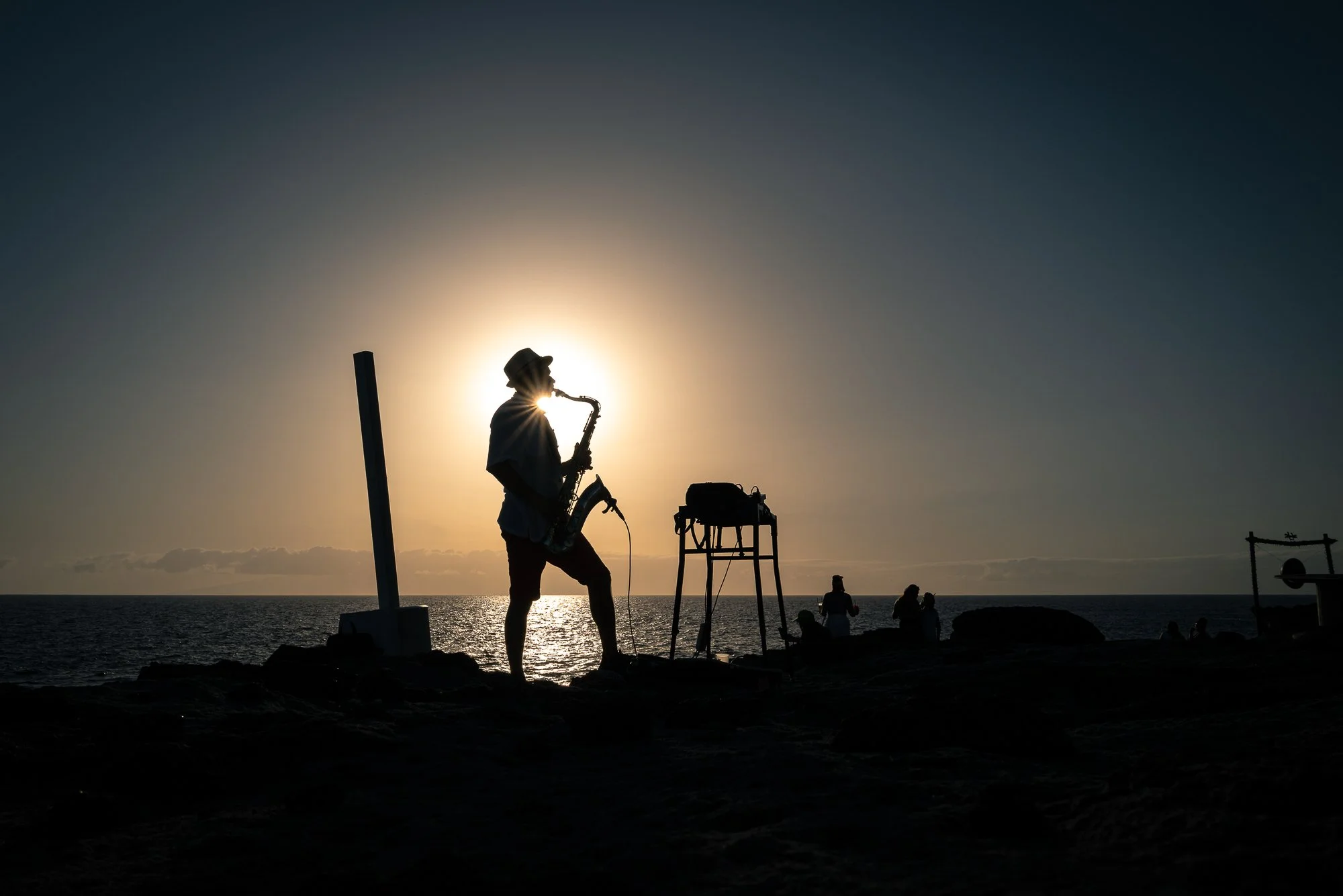A silhouette of a person playing a saxophone on a beach during sunset, with other people in the background near the ocean.