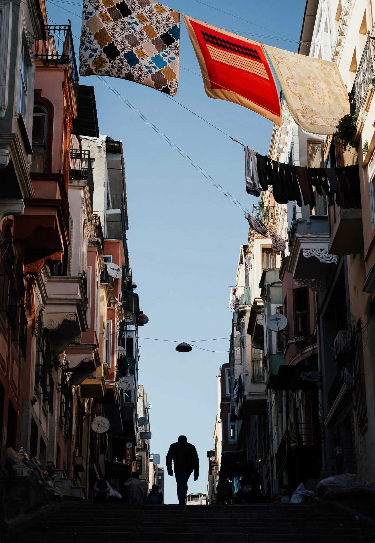 A silhouette of a man walking up stairs in a European city alleyway, with colorful multi-story buildings on either side, laundry hanging above, and clear blue sky overhead.