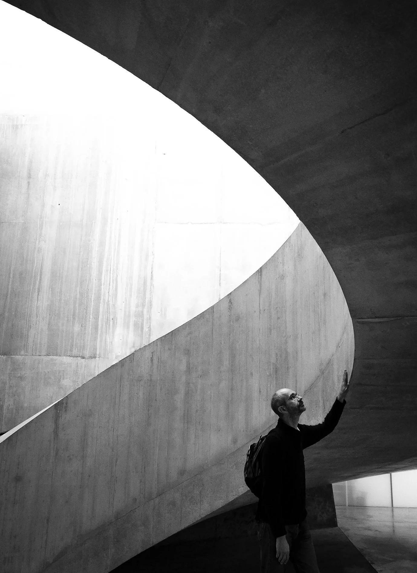 A man with a backpack gesturing upward in an architecturally modern, curved concrete space with geometric shapes and contrasting light and shadow.
