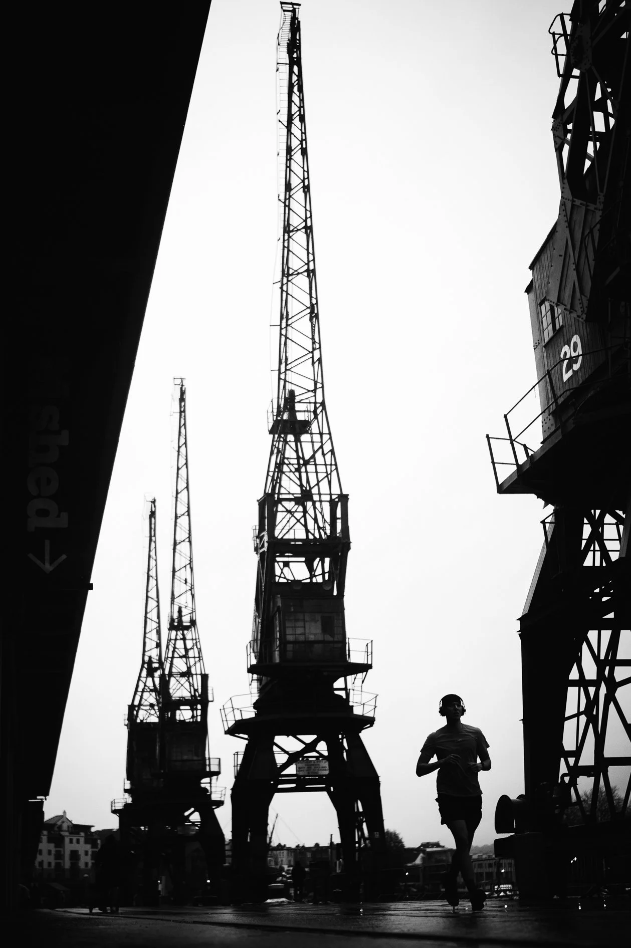 Black and white photo of a woman jogging near construction cranes, with tall structures and a building in the background.