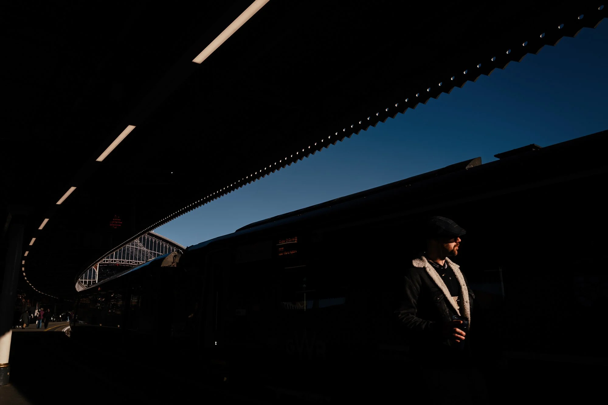 A man wearing a jacket and holding a coffee cup, standing at a bus stop under a large, overhanging roof with lights along the edge, during daytime with clear blue sky in the background.