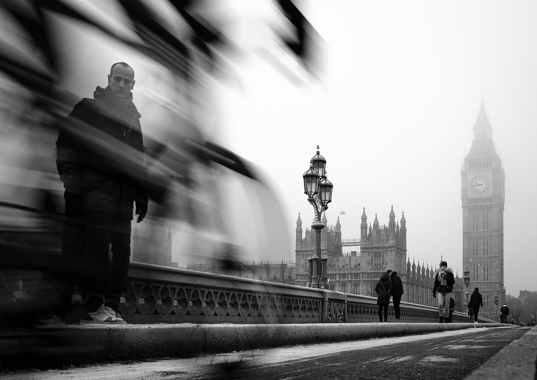 Black and white photo of a man standing on Westminster Bridge with Big Ben in the background, along with some pedestrians and traditional street lamps.