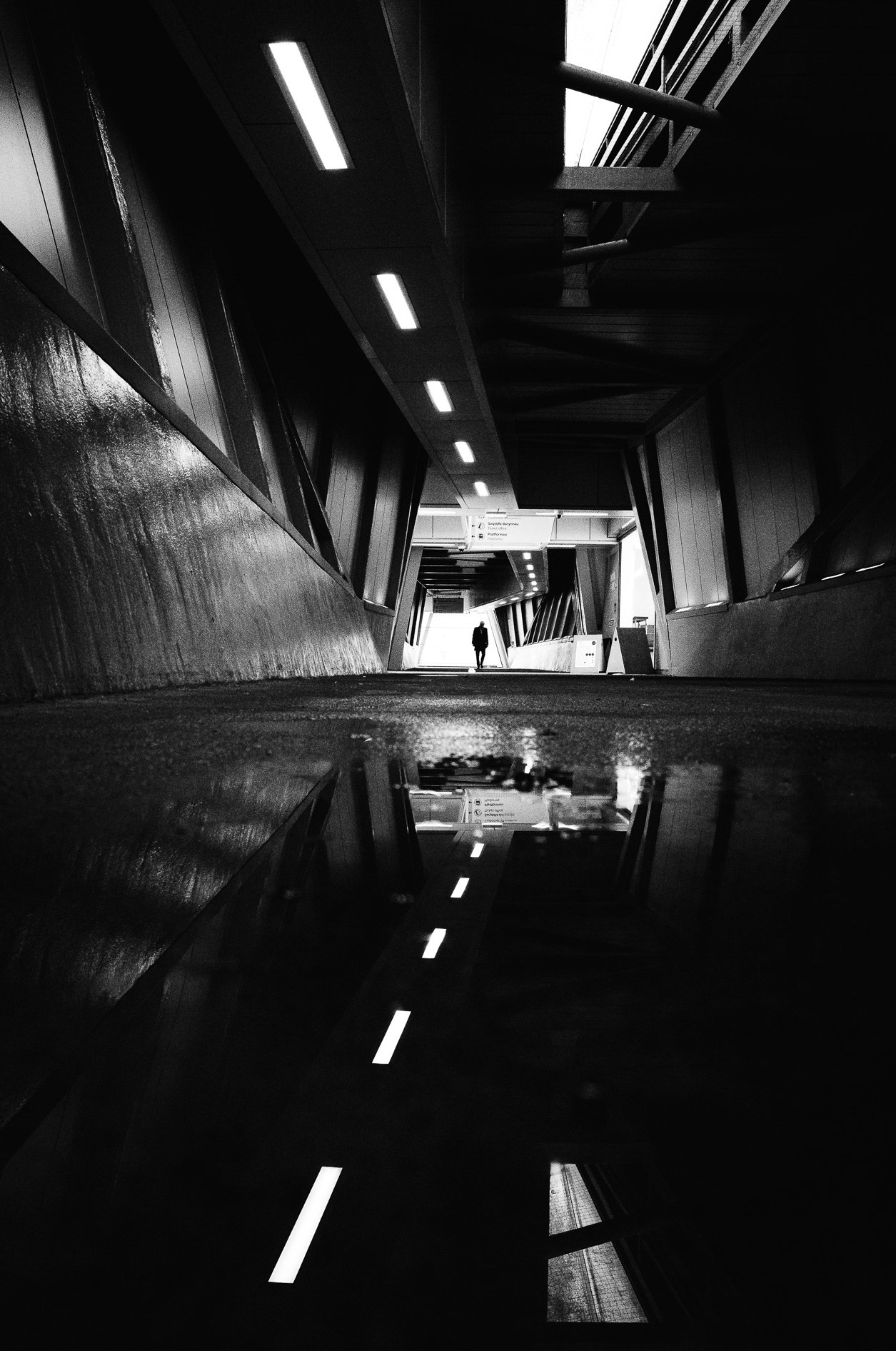 Black and white photo of an indoor corridor with a person walking towards a lighted exit at the end, reflecting off the shiny floor, with ceiling lights and modern architecture