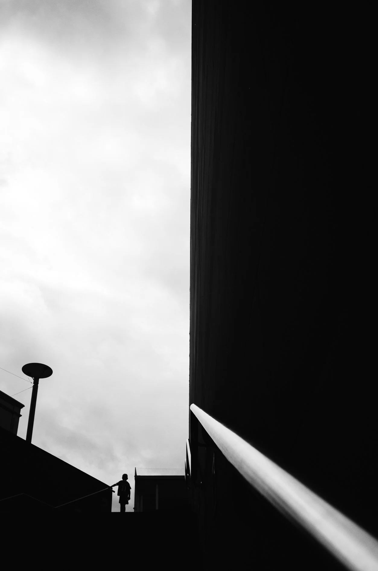 Black and white photo of a street scene with a person walking, a streetlamp, a large dark building, and cloudy sky.