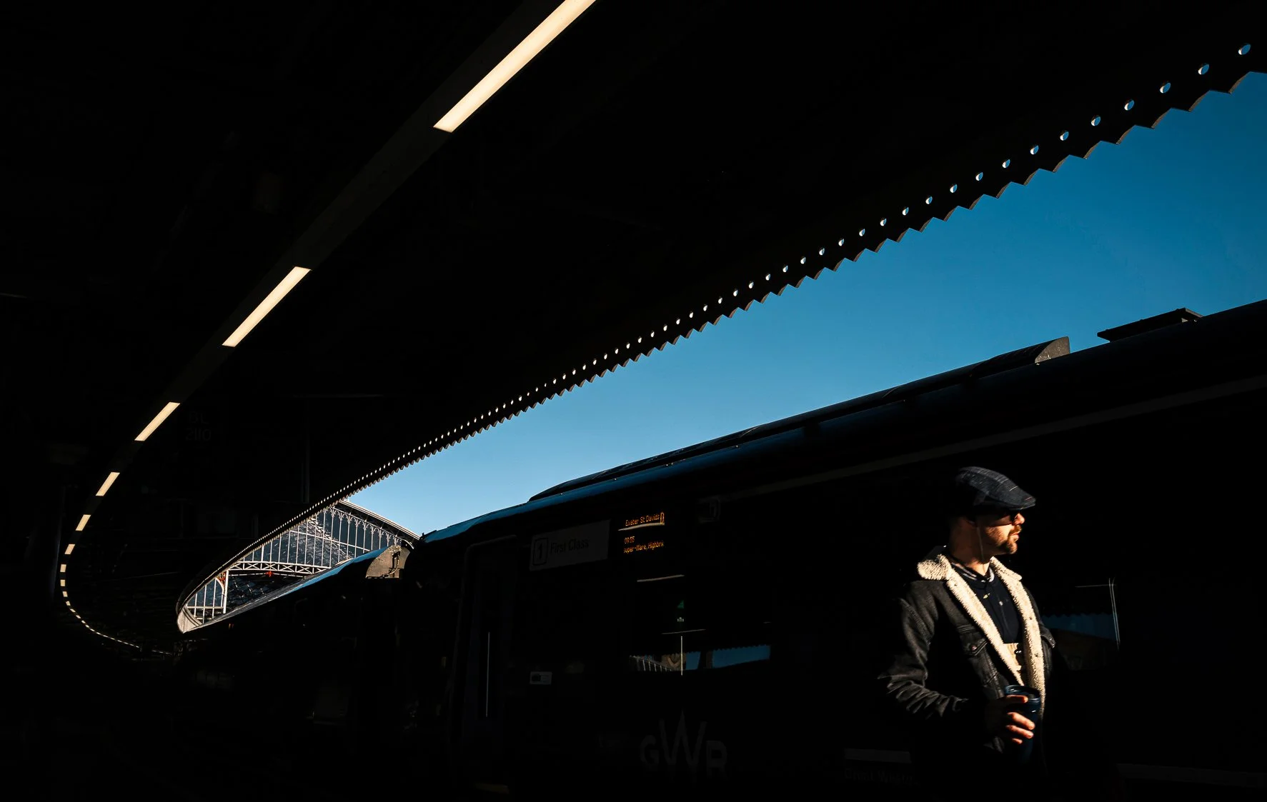 A person with a beard wearing a cap and sunglasses holding a drink at a bus stop under a dark canopy, with sunlight illuminating part of the scene against a blue sky.