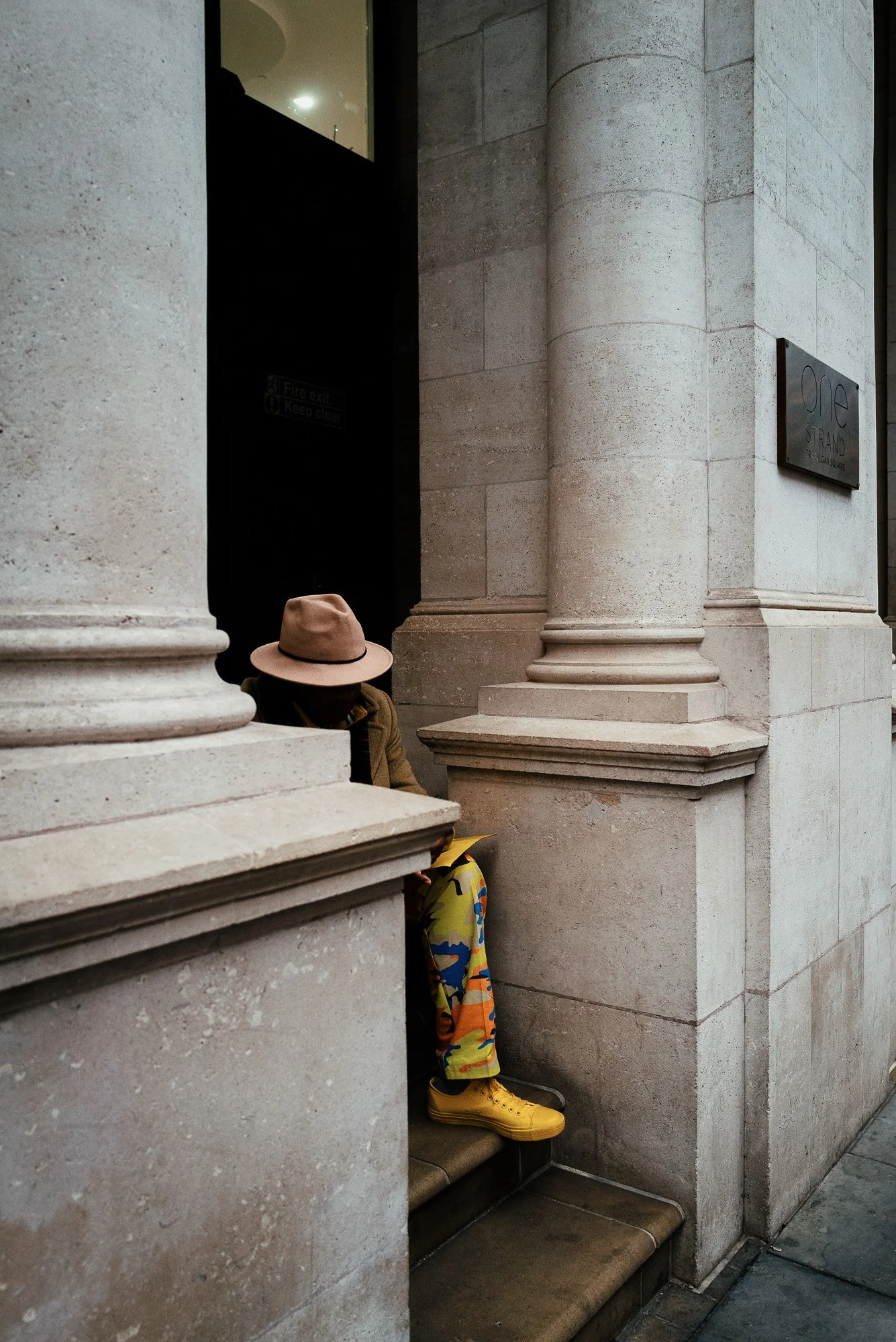 A person wearing a beige hat, camouflage pants, and yellow shoes is sitting behind a large stone column on steps outside a building. The person's face is hidden behind the column and hat.