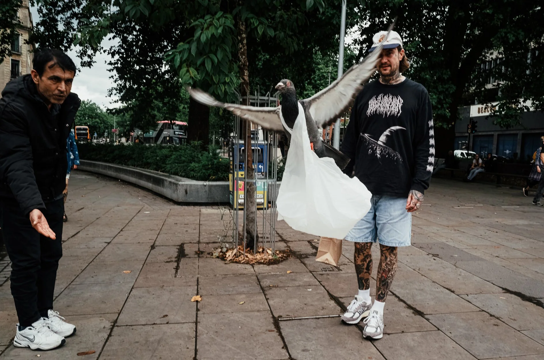 A man with tattoos on his legs and wearing a black shirt with graphic art, carrying a large bird sculpture, stands next to another man in dark clothing on a city sidewalk with trees and buildings in the background.