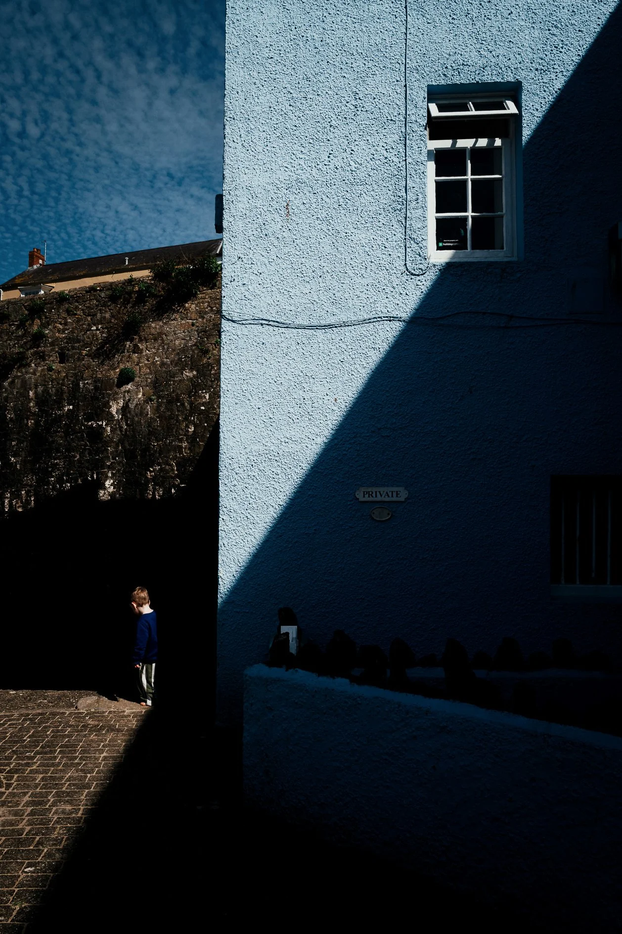 A light blue textured building wall with a window, partially shaded by shadow, and a sign that reads 'PRIVATE'. In the foreground, a only part of a person with a blue shirt is visible, with their head down, standing on a cobblestone ground. The sky i