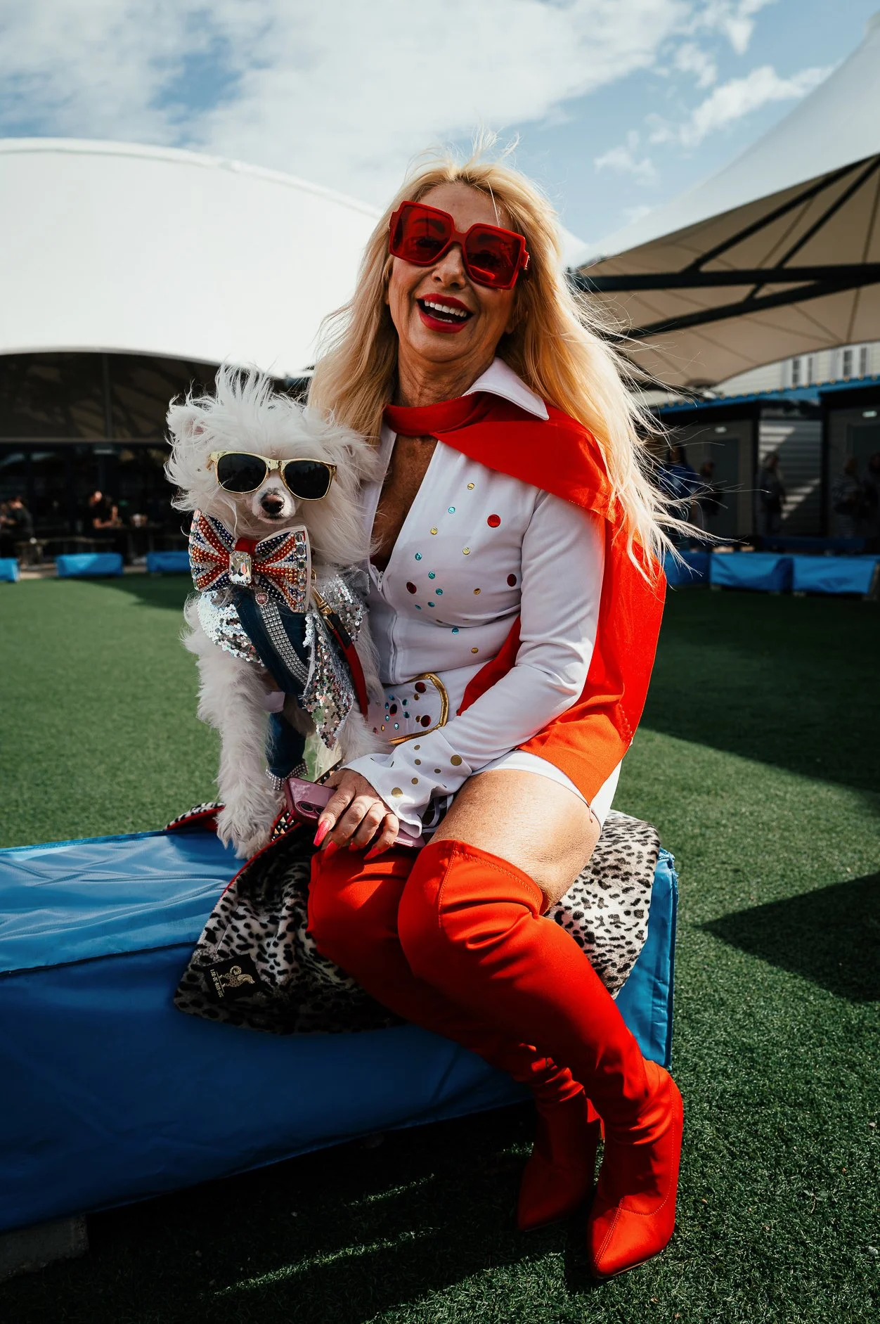 A joyful woman with blonde hair wearing red sunglasses, a white and red outfit with colorful polka dots, and bright red over-the-knee boots, sitting outdoors on a blue cushioned lounge chair. She is holding a small white dog wearing sunglasses, a seq