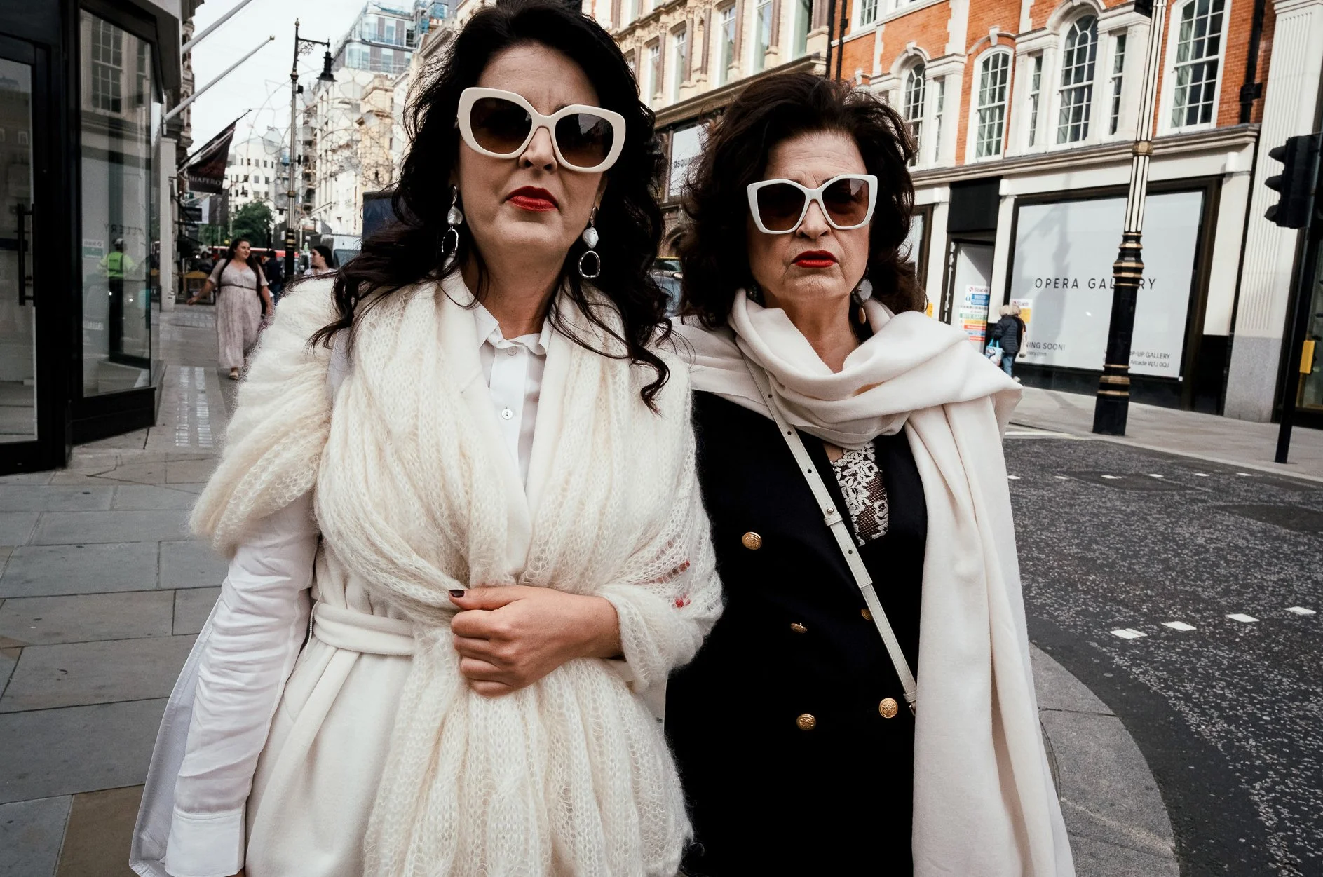 Two women in stylish white outfits and large sunglasses standing on a city street with historic brick buildings, storefronts, and pedestrians in the background.
