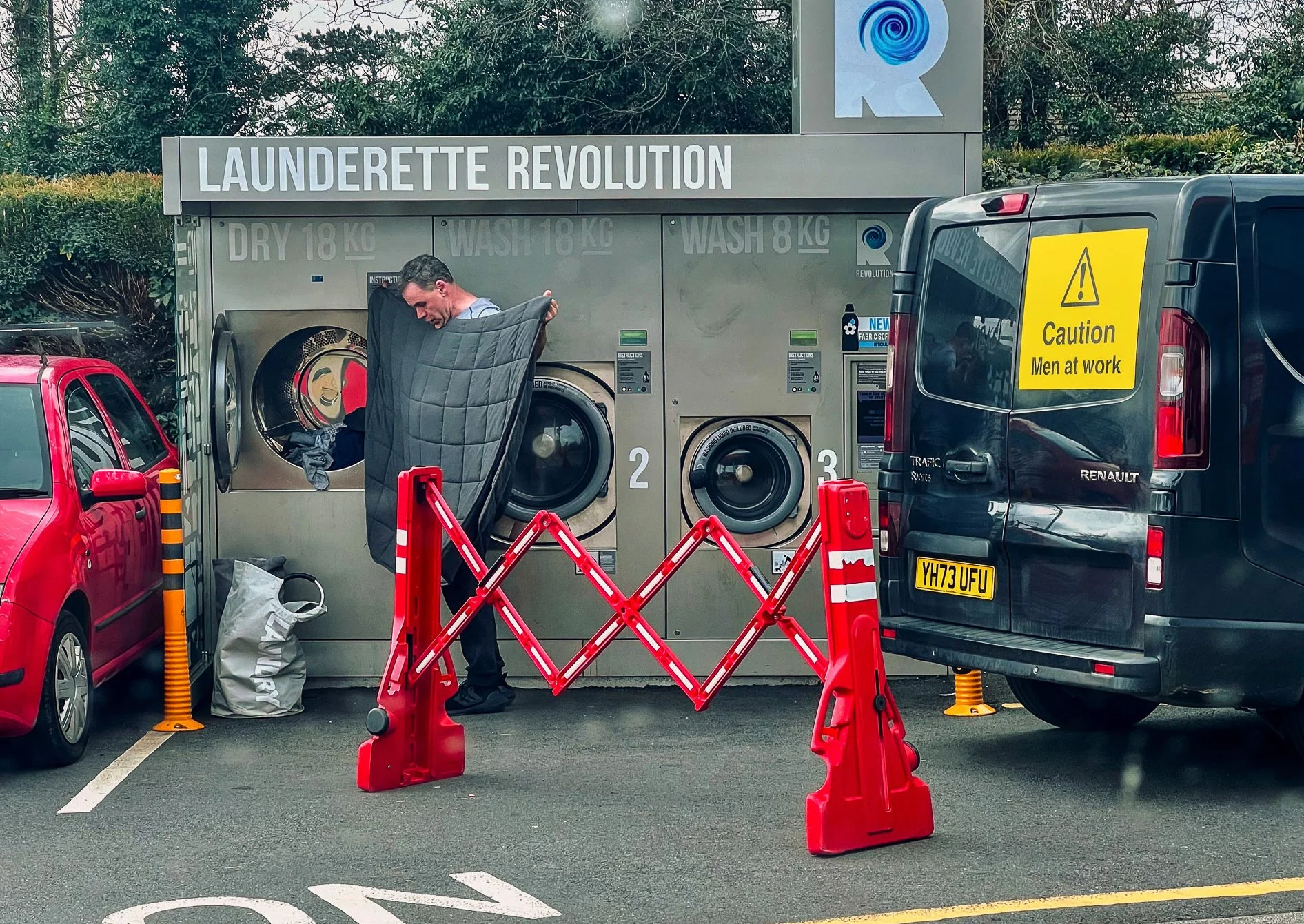 Man folding a quilt at a laundrette. The laundrette has a sign that says 'LAUNDERETTE REVOLUTION' with instructions for dry and wash cycles. There are two washing machines and a black van parked nearby with a yellow caution sign that says 'Caution Me