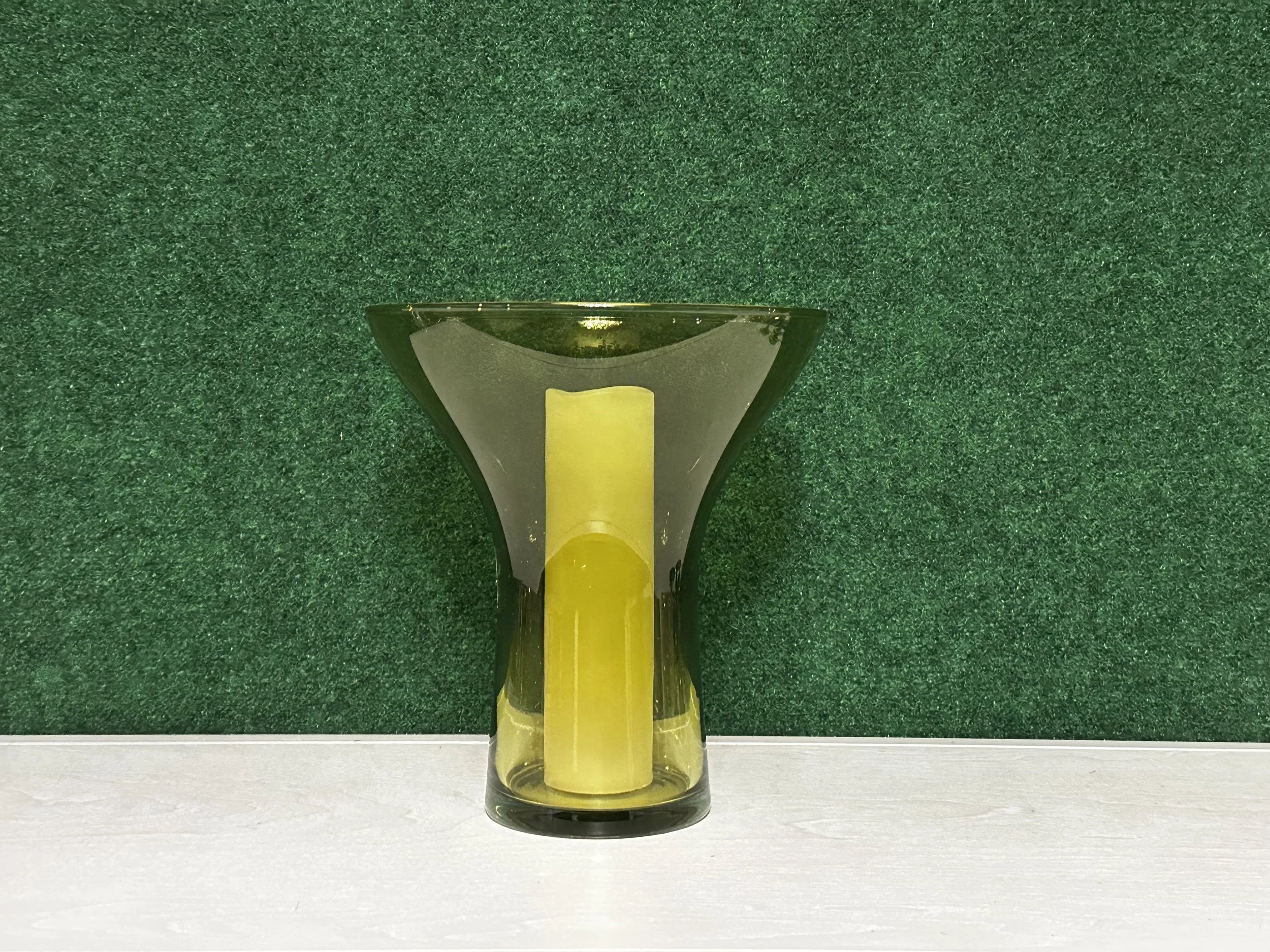 Green glass vase with a yellow candle inside, set against a green textured background.