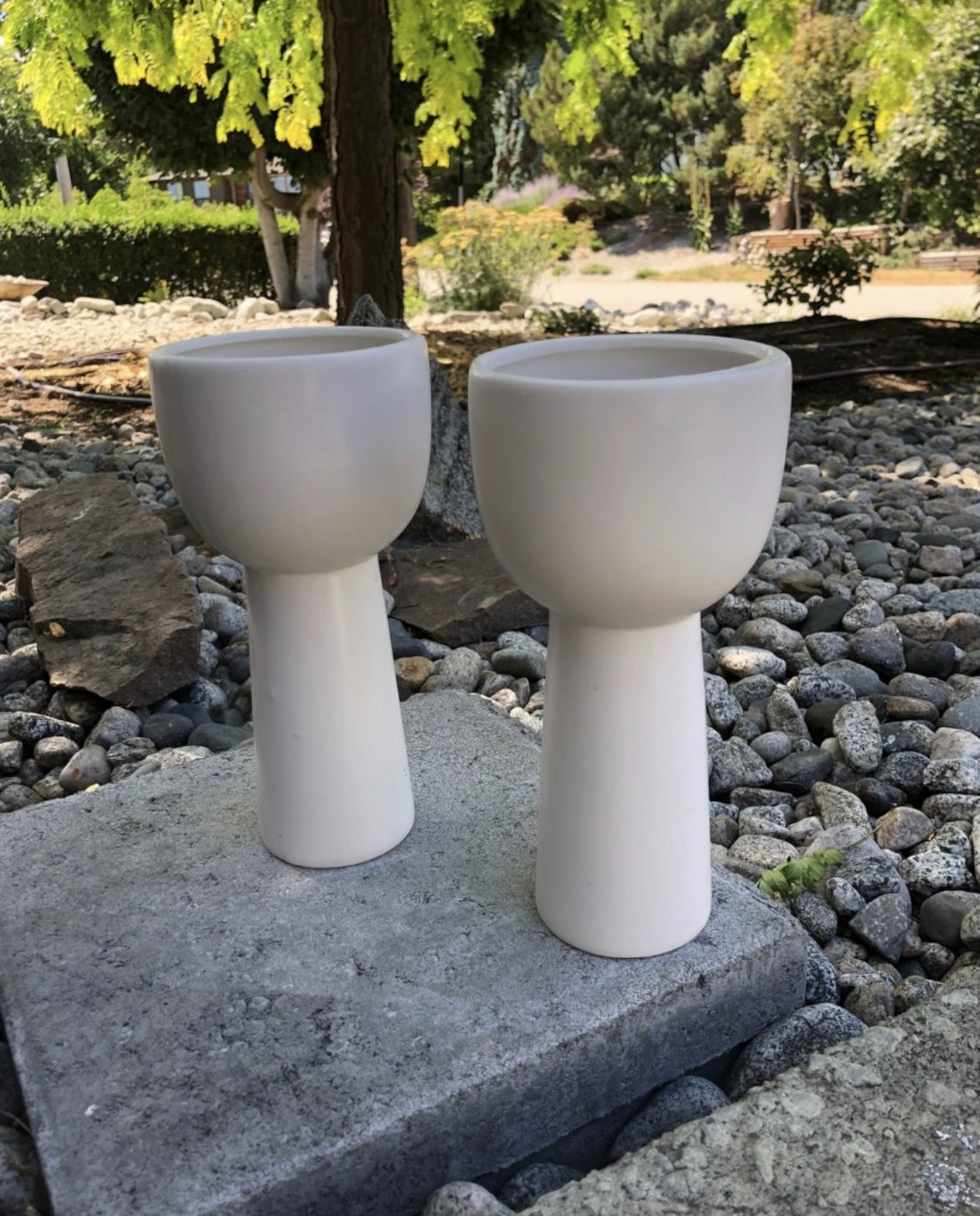 Two tall white ceramic planters on a stone surface with rocks and greenery in the background.
