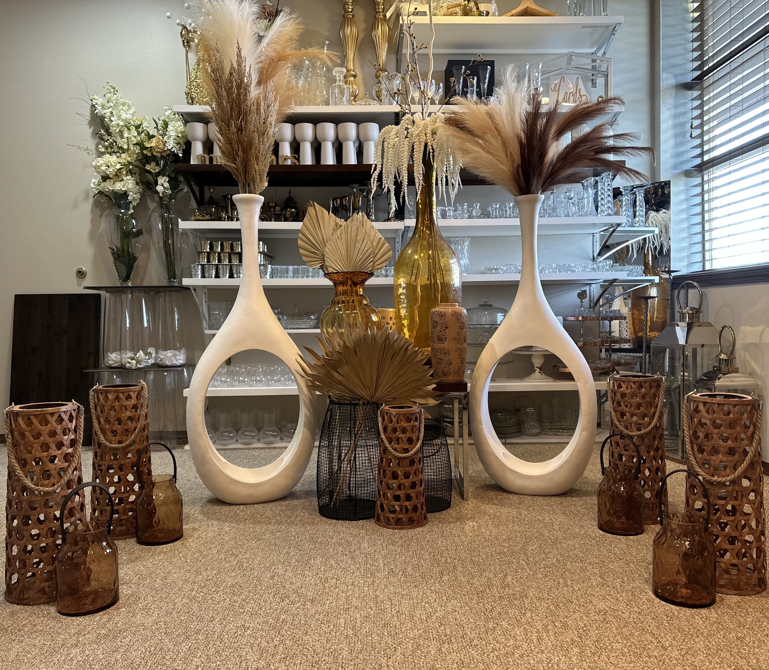 Interior display with decorative vases, glassware, and wicker lanterns on a carpeted floor. Tall white vases with pampas grass, amber glass bottles, and dried palm leaves are arranged in the foreground. Shelves in the background hold various glass and ceramic items.