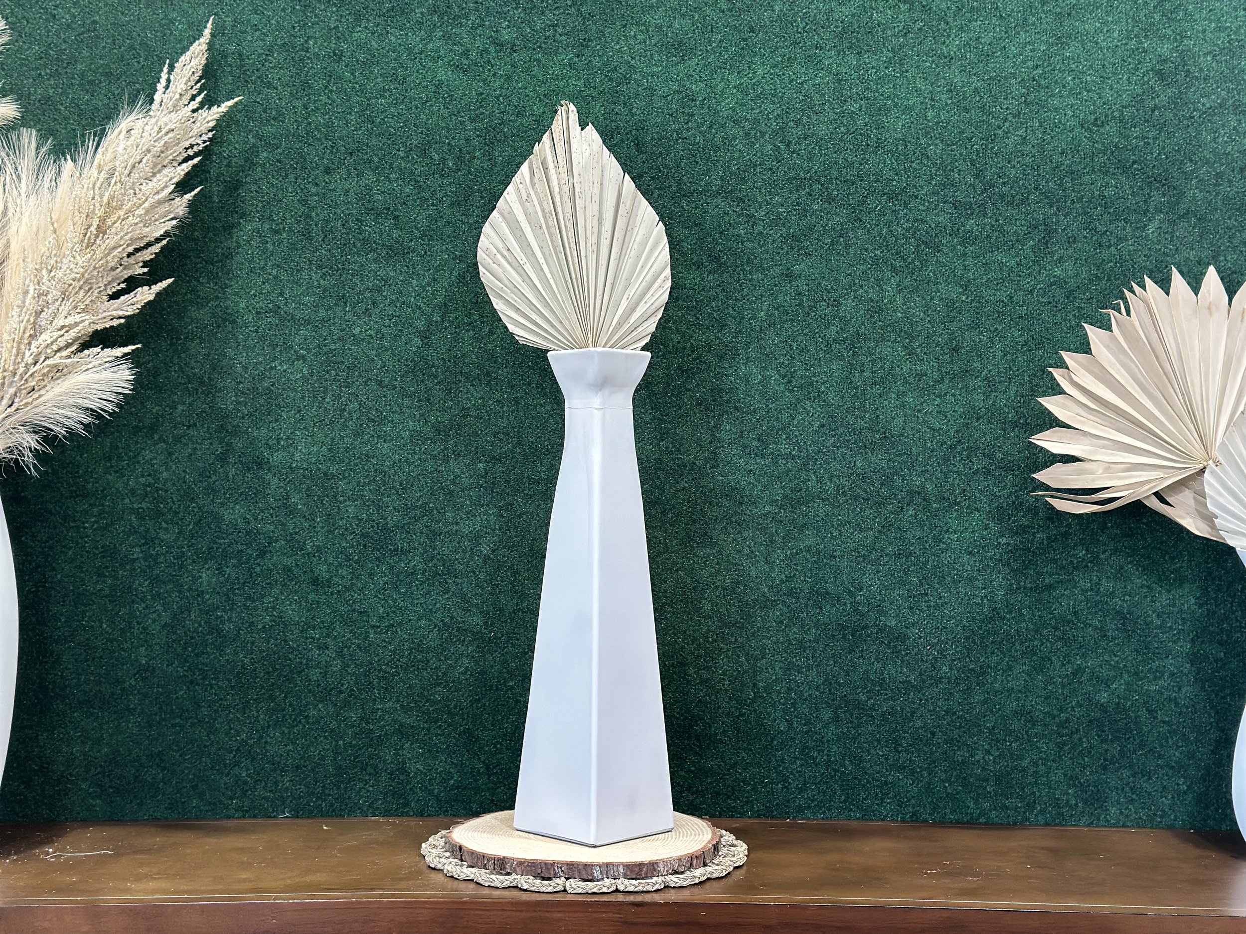 Minimalist white vase with geometric design holding a dried palm leaf centerpiece, flanked by pampas grass and dried palm leaves, on a dark green background.