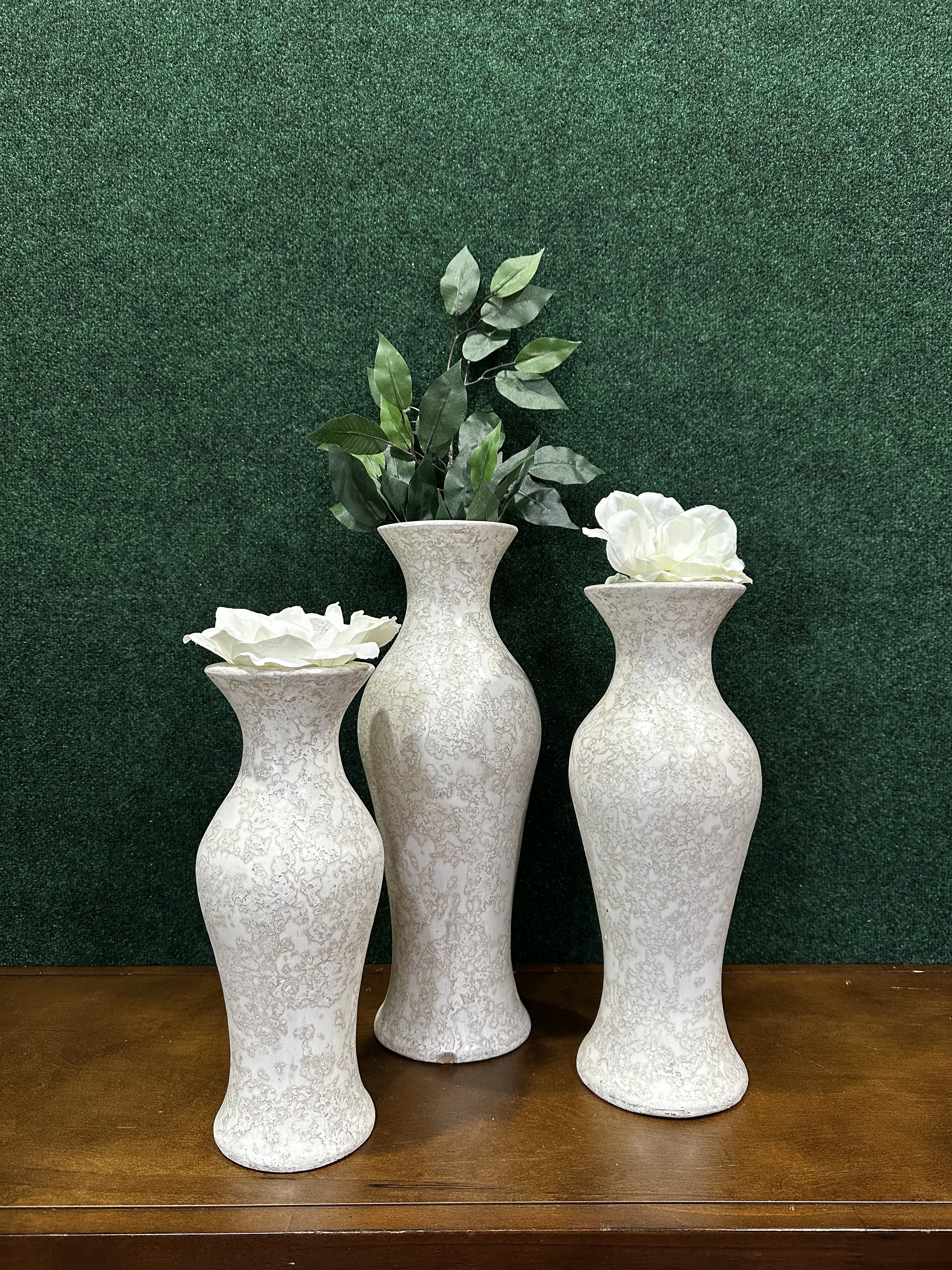 Three decorative vases with white roses and green leaves on a wooden surface against a green backdrop.