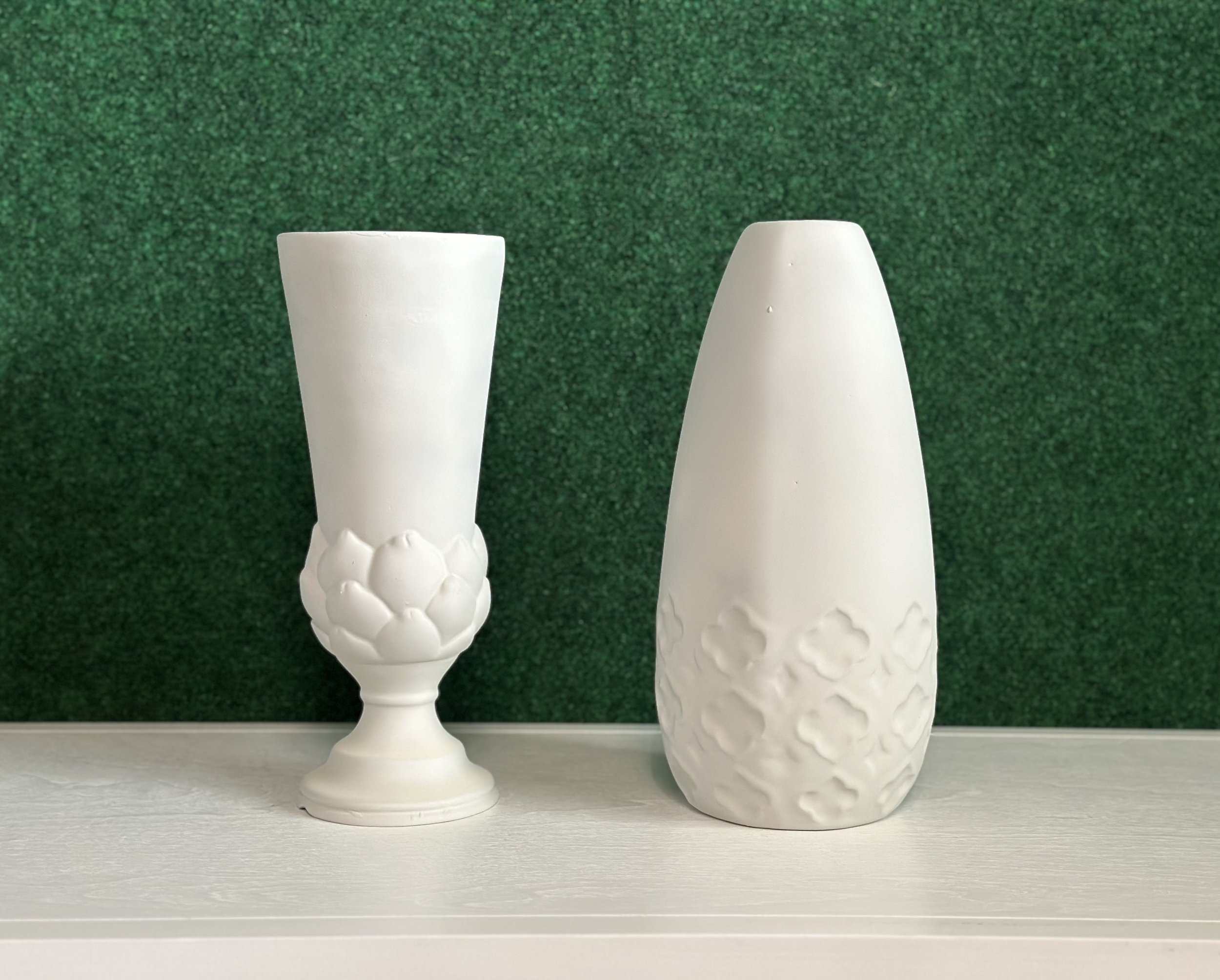 Two white ceramic vases with textured designs on a light surface against a green background.