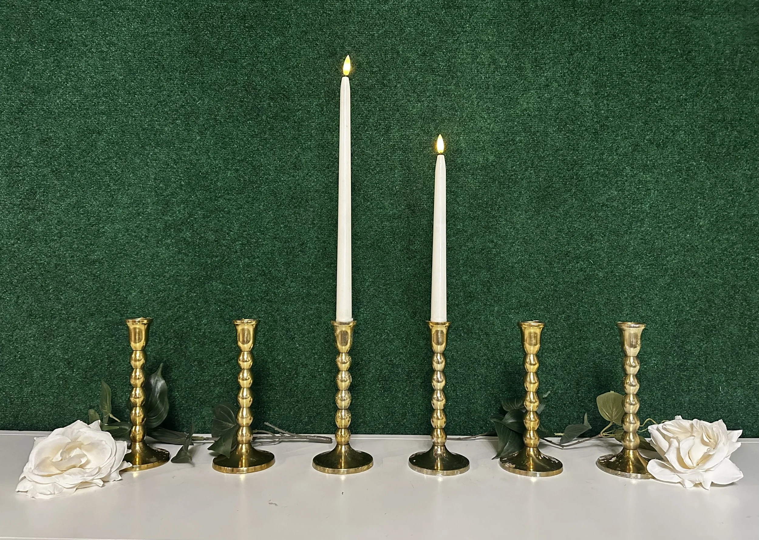Six brass candlesticks on a white surface with two white lighted taper candles in the middle; fake white roses and green leaves are placed on each end. Green background.