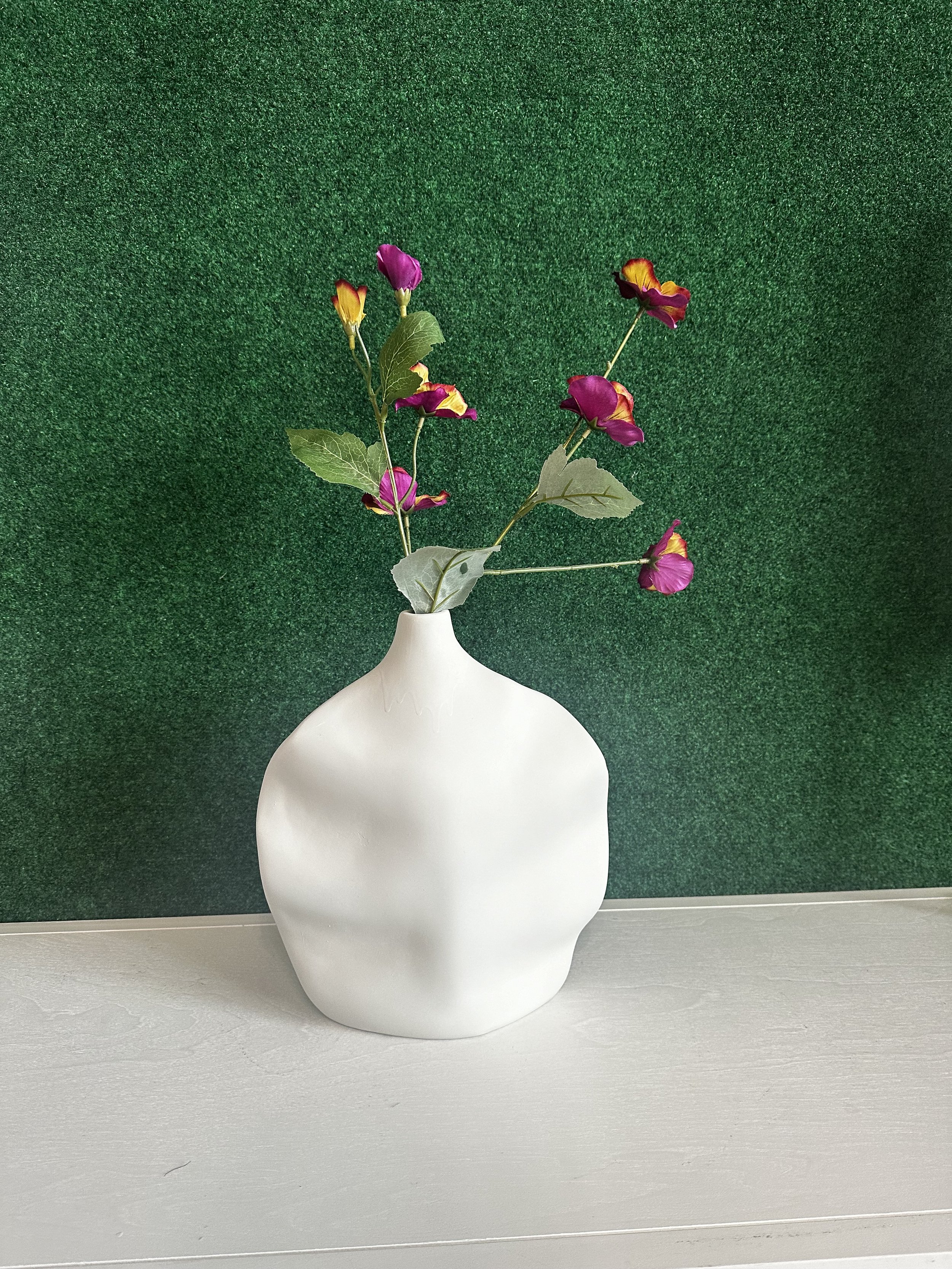 White vase with abstract shape holding purple and yellow artificial flowers against a green textured background.