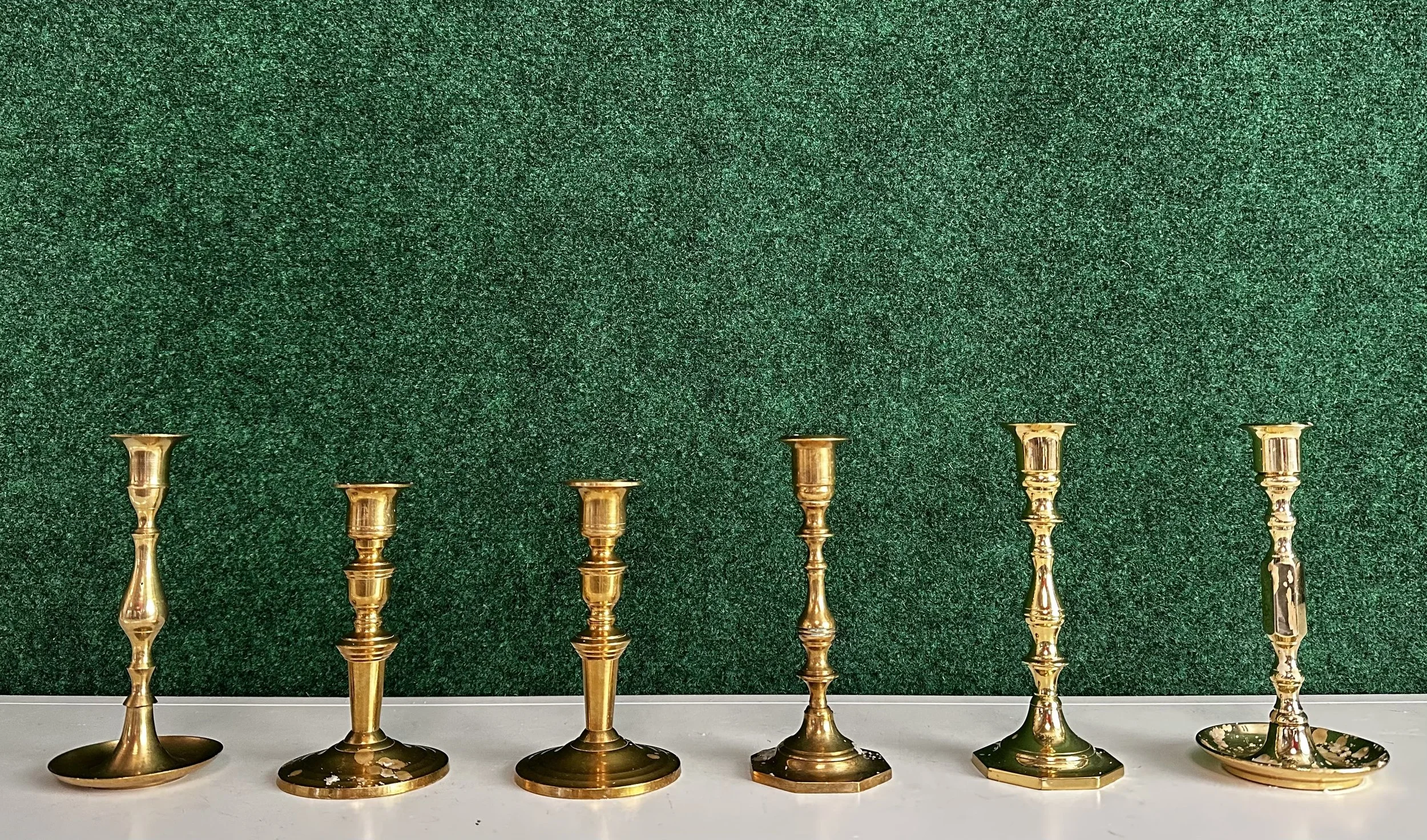 Six brass candlesticks arranged in a row on a white surface against a green background.