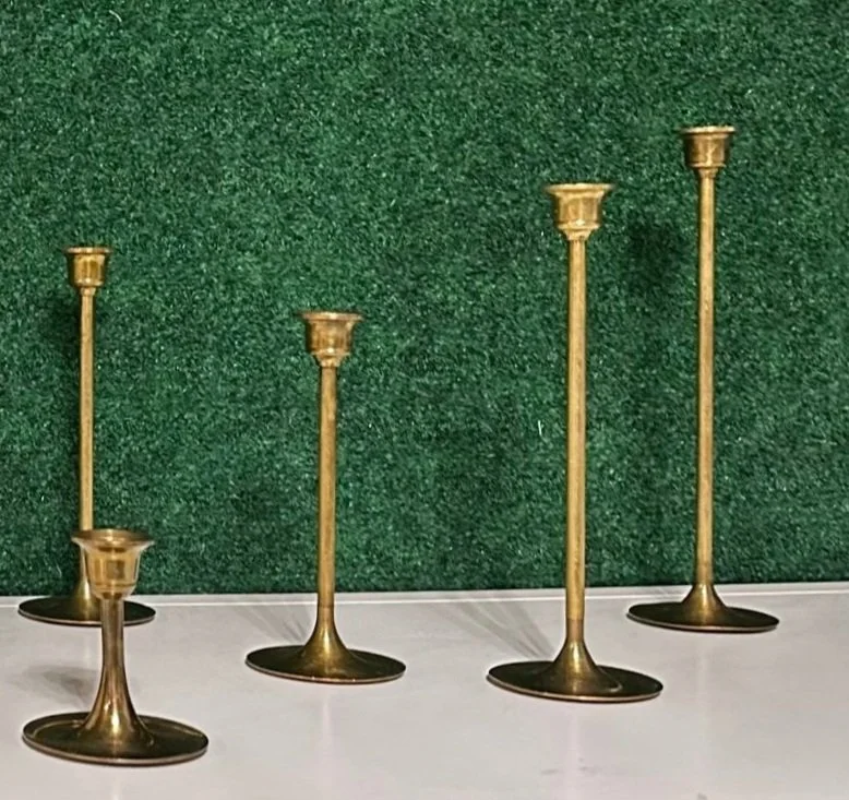 Five brass candle holders of varying heights on a white surface against a green background.