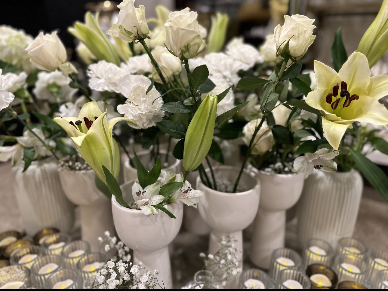 White flowers and lilies in ceramic vases surrounded by small candle holders.