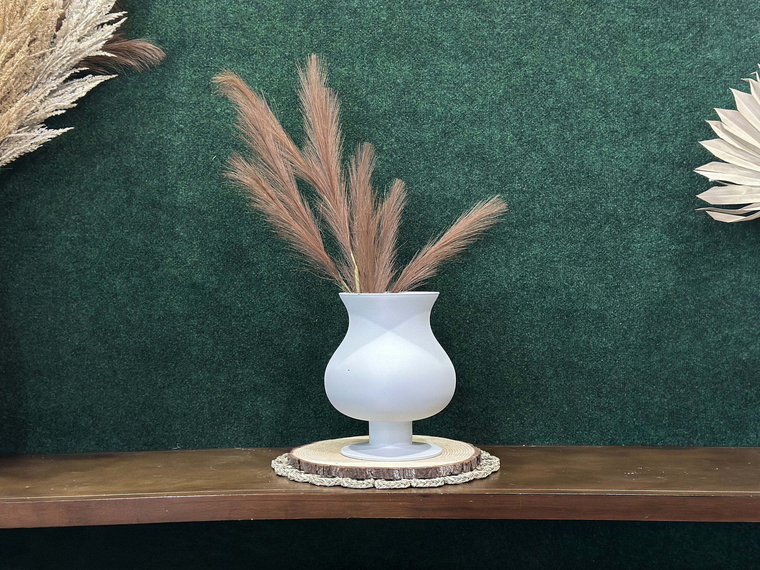 A white vase with dried pampas grass on a wooden shelf, set against a dark green backdrop.