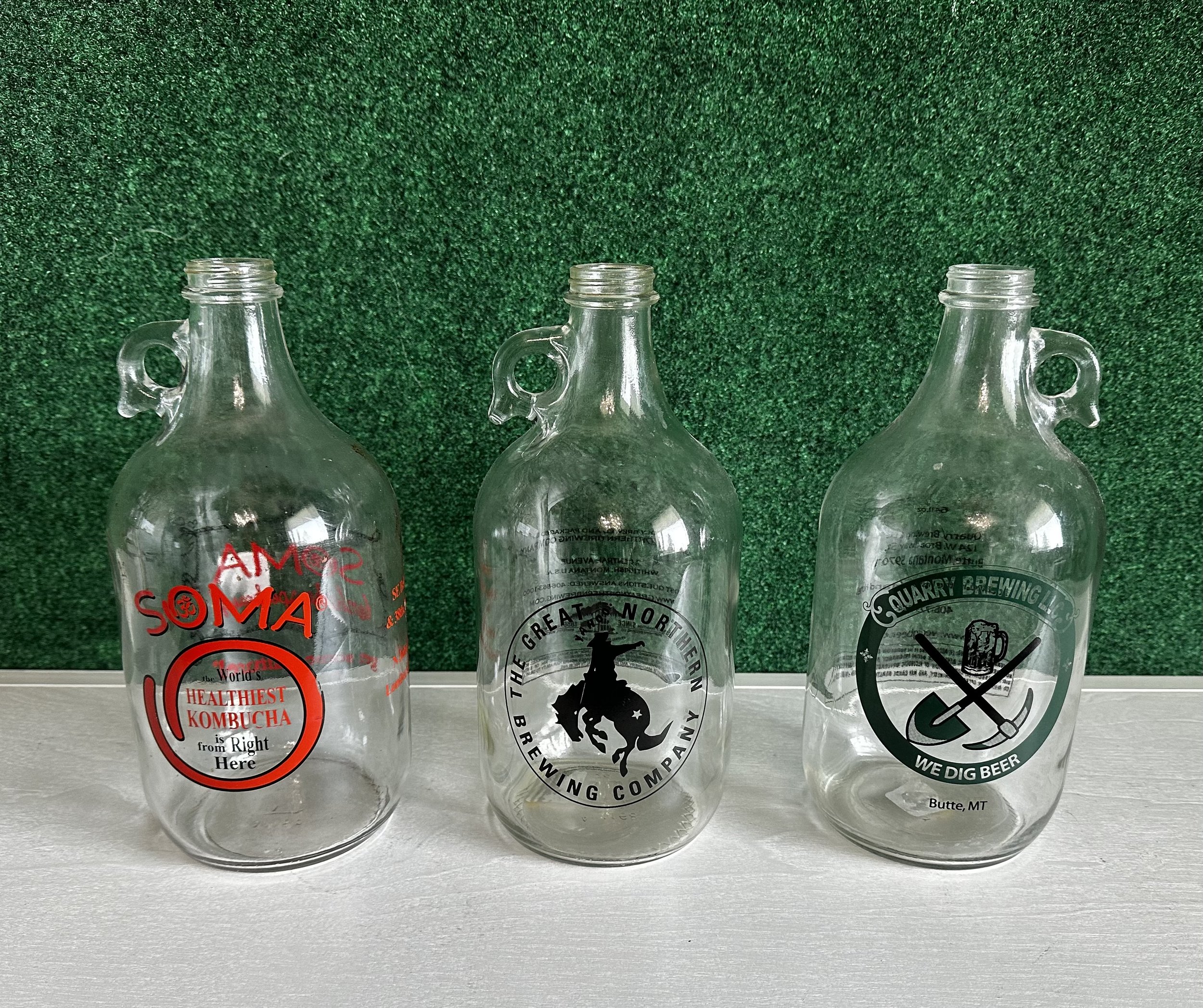 Three empty glass growlers with handles, each featuring different brewery logos and text designs, placed in front of a green background.