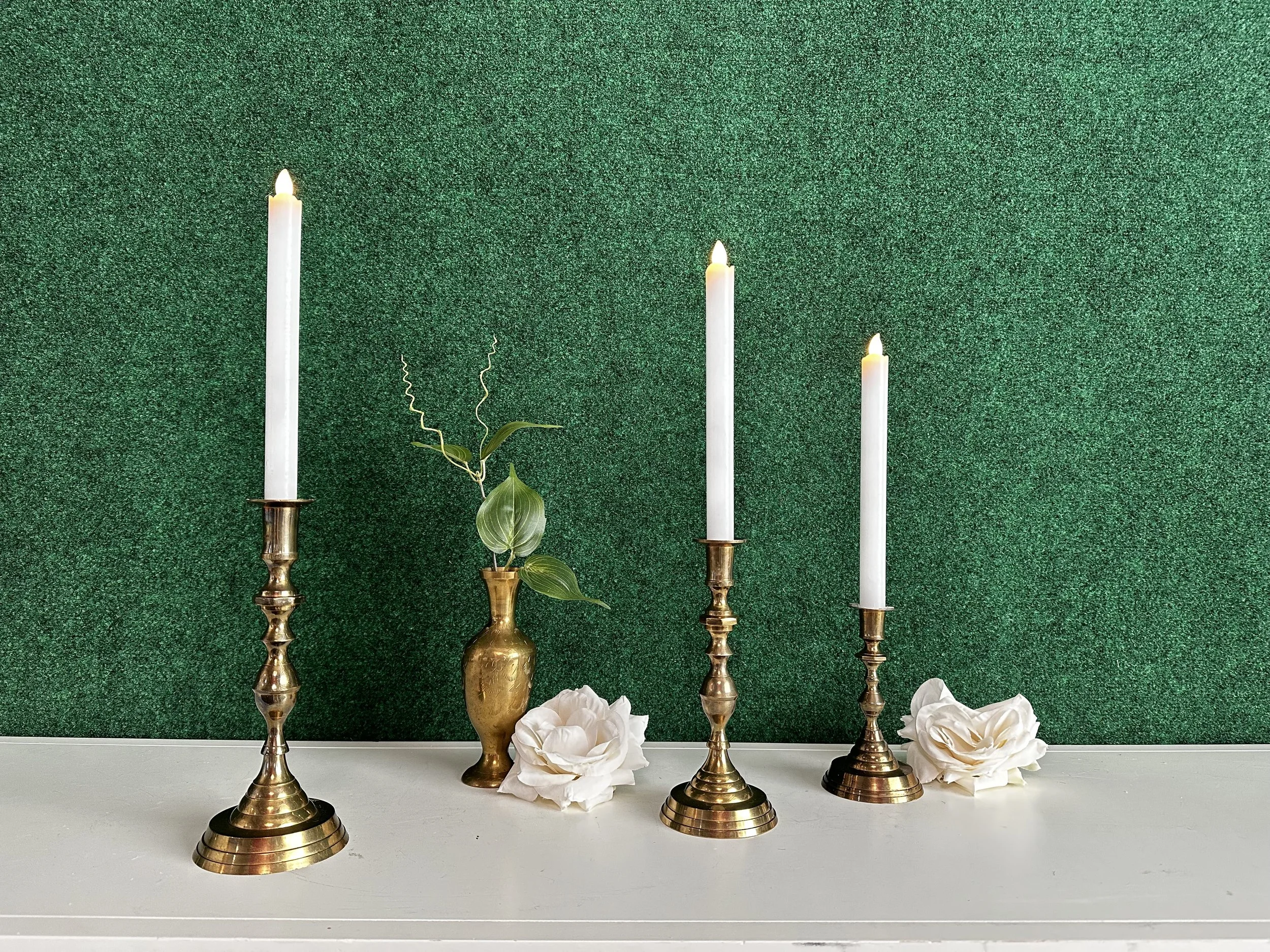 Three brass candlesticks with lit white candles and a brass vase with green leaves on a white surface, set against a green textured background with white rose decorations.