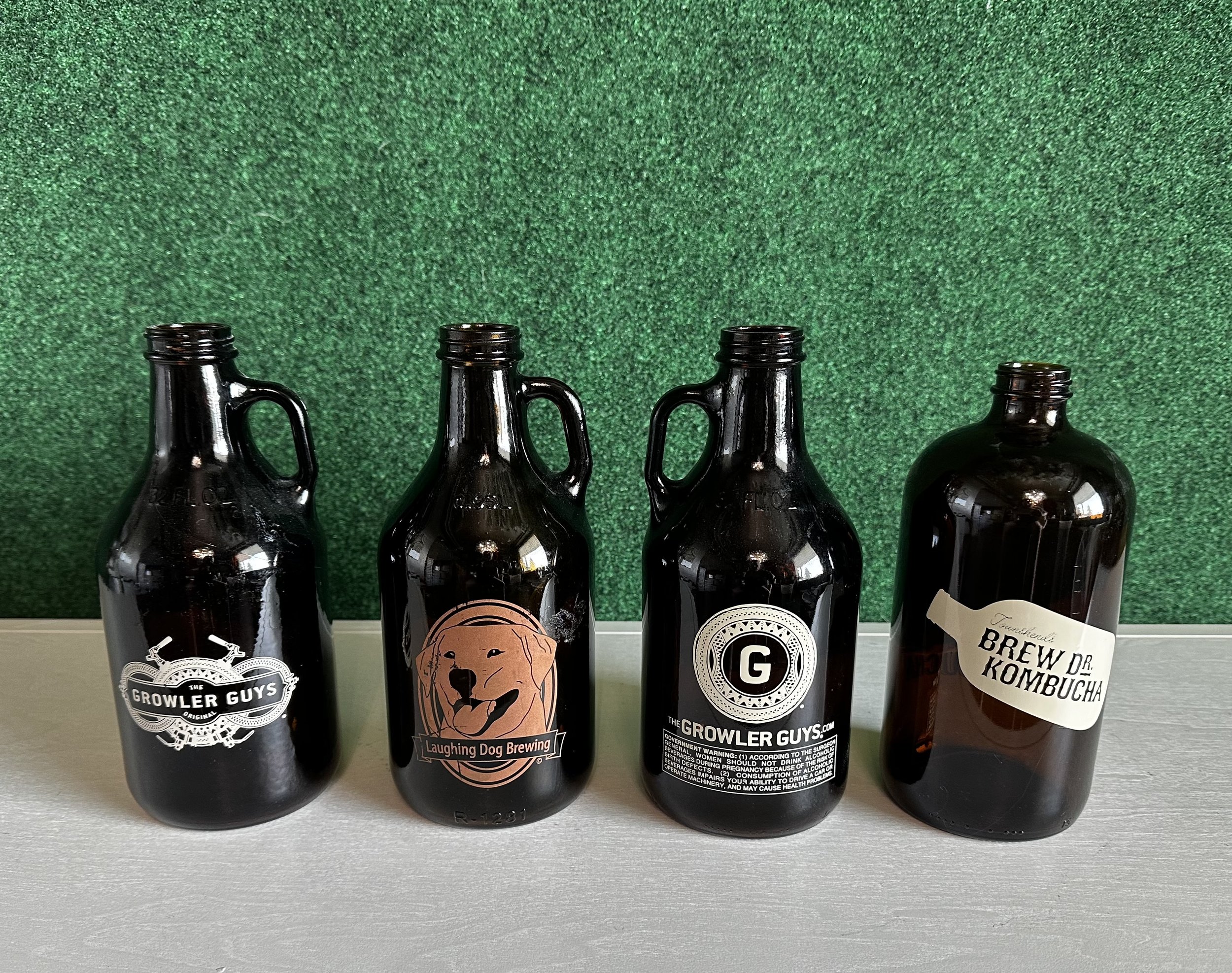 Four dark glass growlers on a surface, each with a different label; two labeled "Growler Guys," one labeled "Laughing Dog Brewing," and one labeled "Brew Dr. Kombucha."
