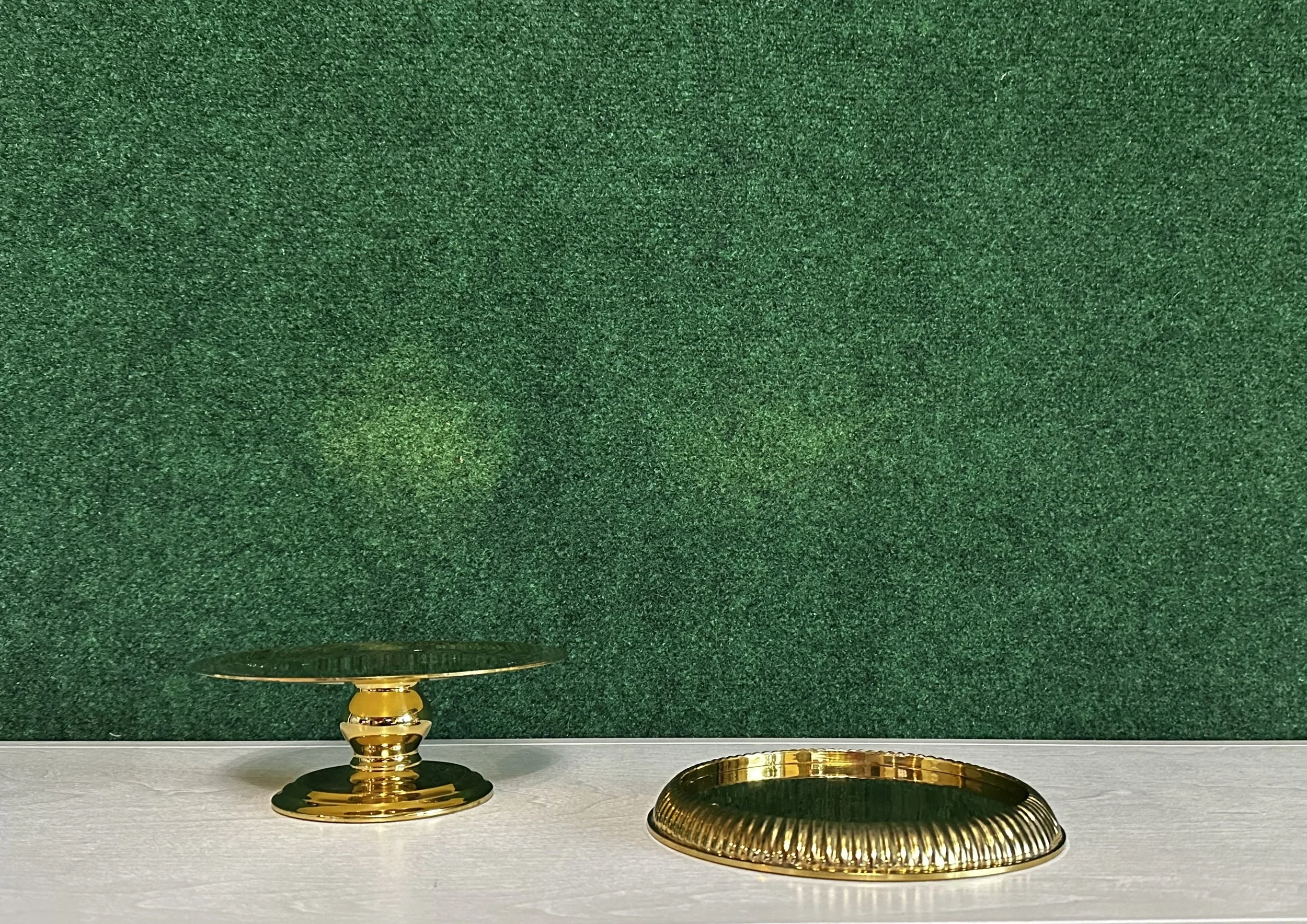 Gold pedestal stand and gold decorative tray on white surface against green background.