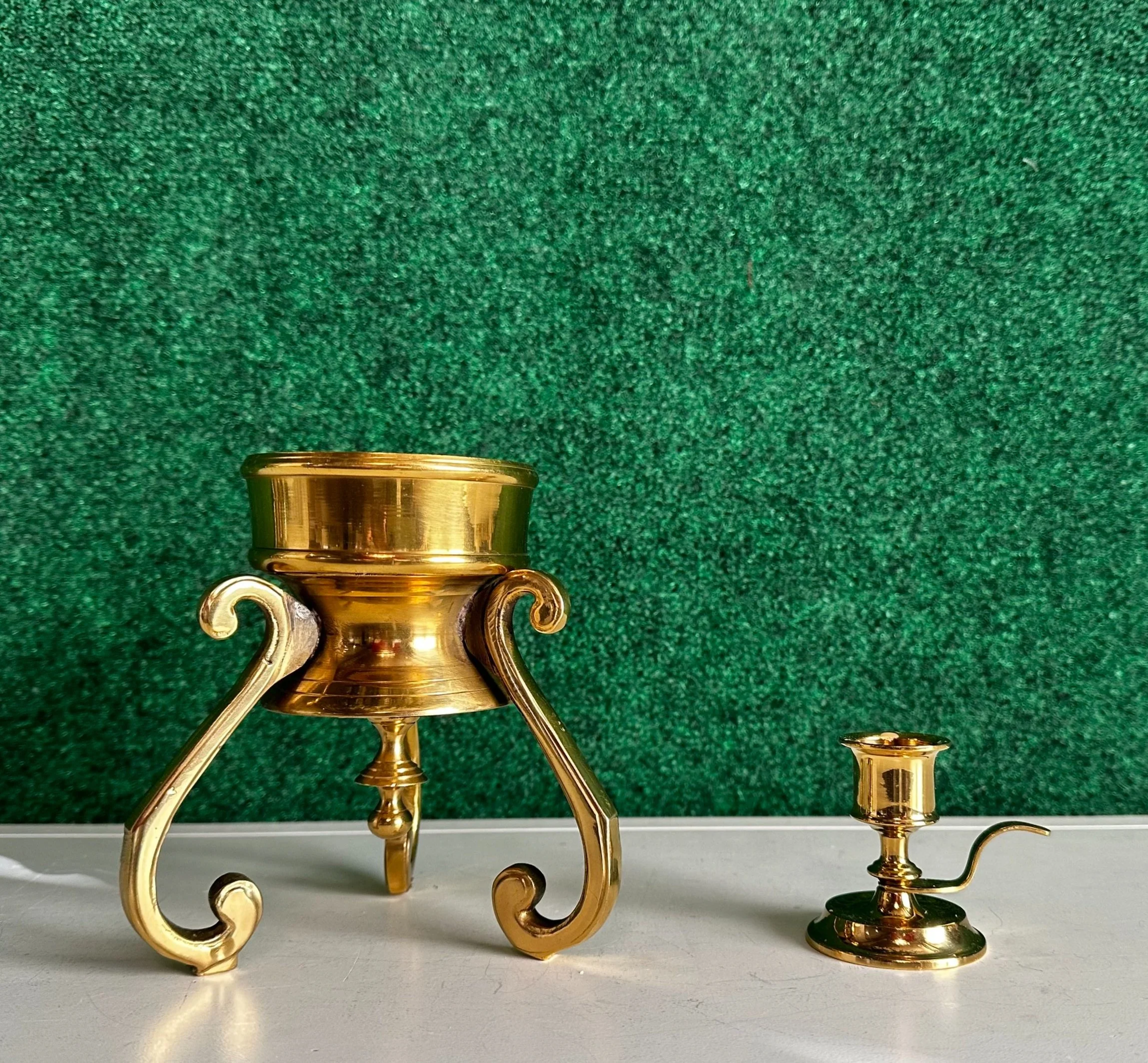 A gold-colored brass candle holder with ornate legs and a small, separate candle snuffer on a textured green background.