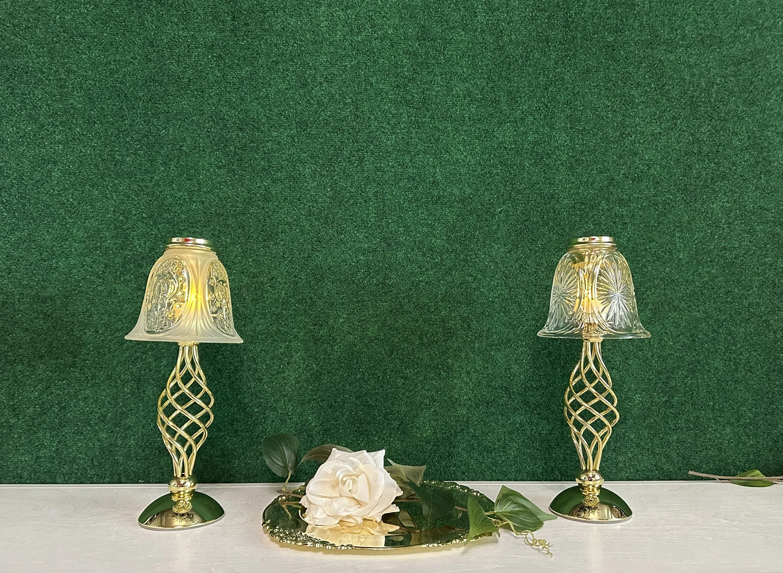 Two decorative gold lamps with intricate glass shades on a green background, accompanied by artificial white flower and green leaves on a mirrored tray.