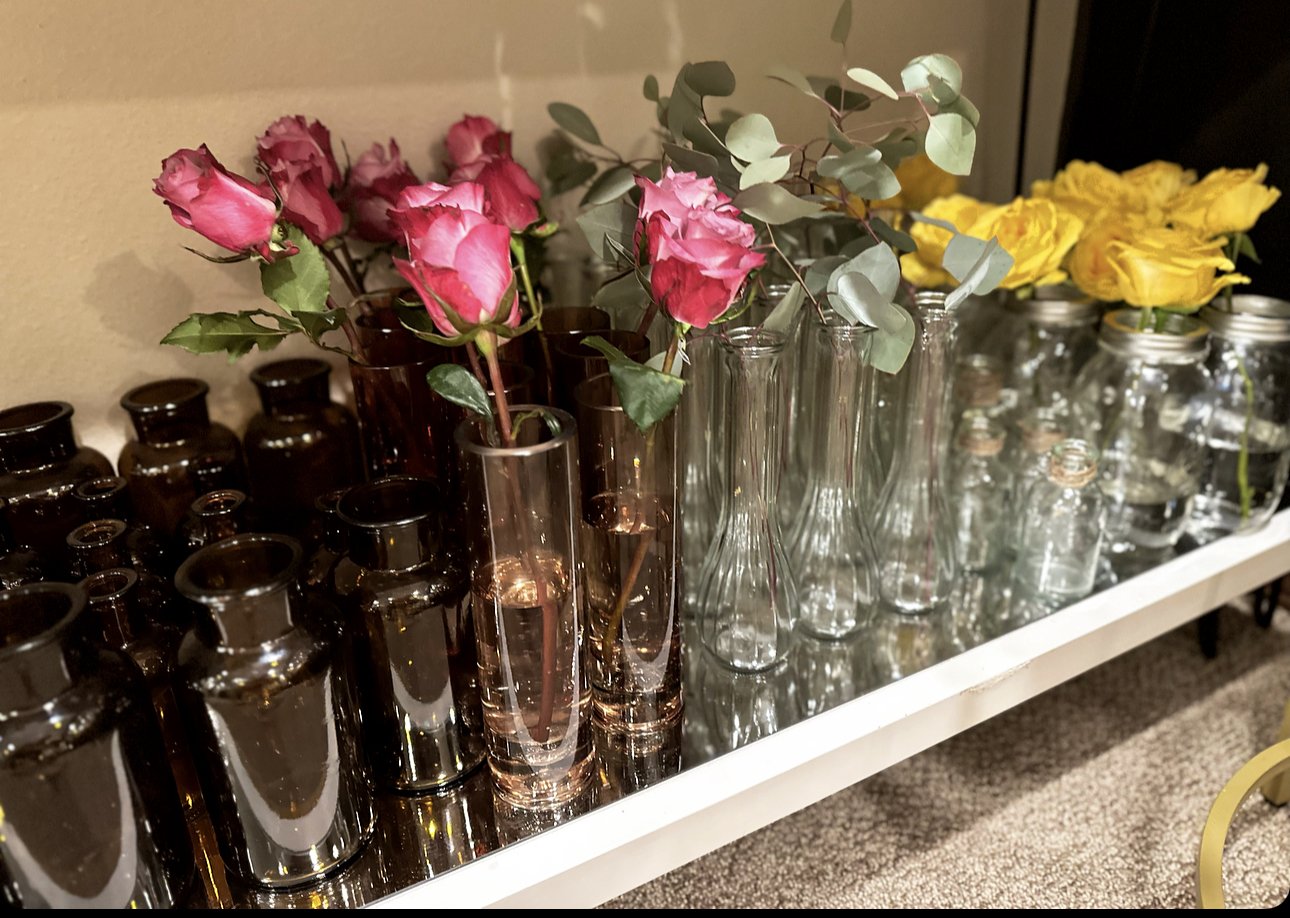 Various vases and bottles on a shelf with pink and yellow roses, and eucalyptus leaves.