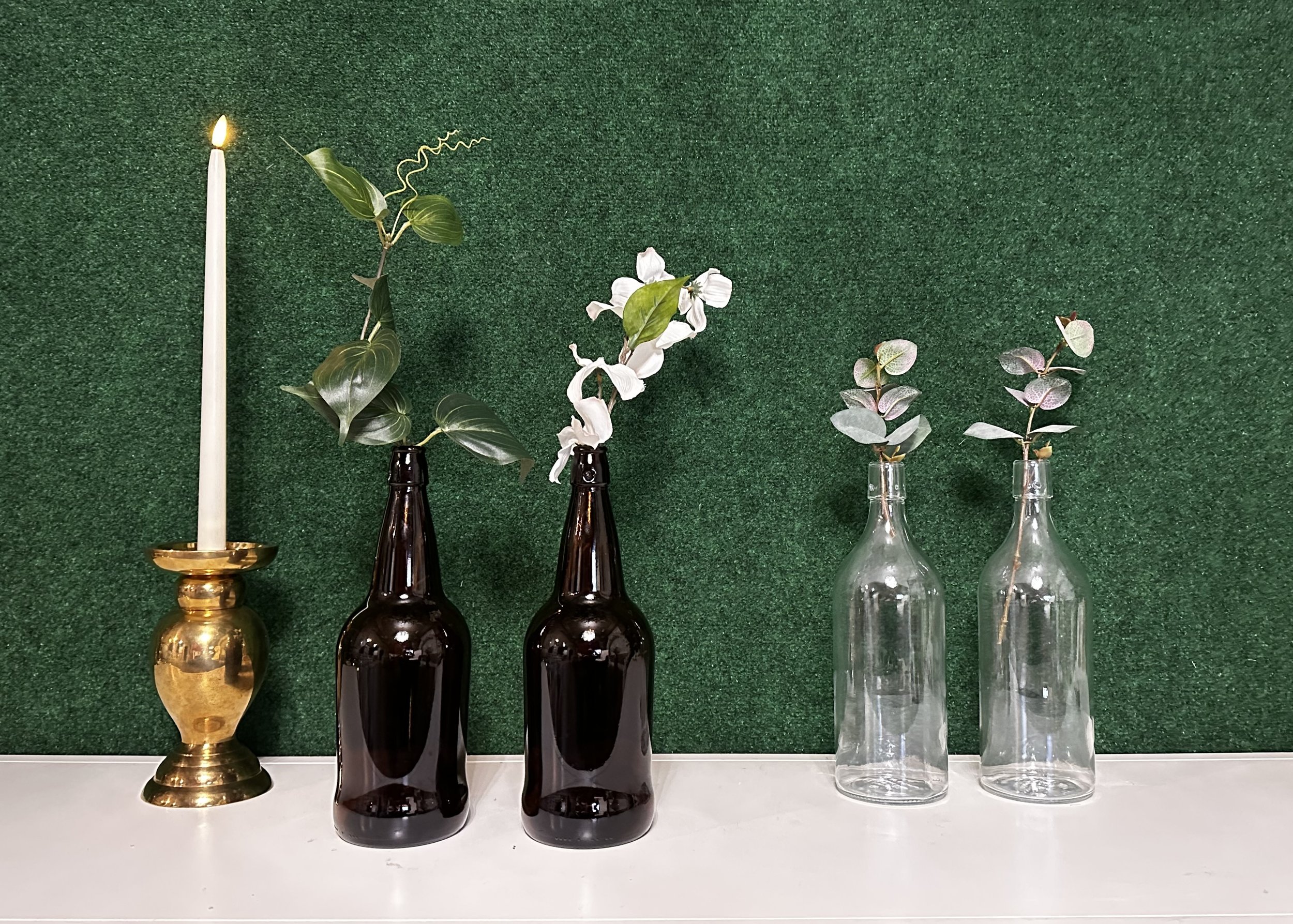 Decorative arrangement featuring a lit candle in a brass holder, brown glass bottles with green and white foliage, and clear glass bottles with small branches, all set against a green textured background.