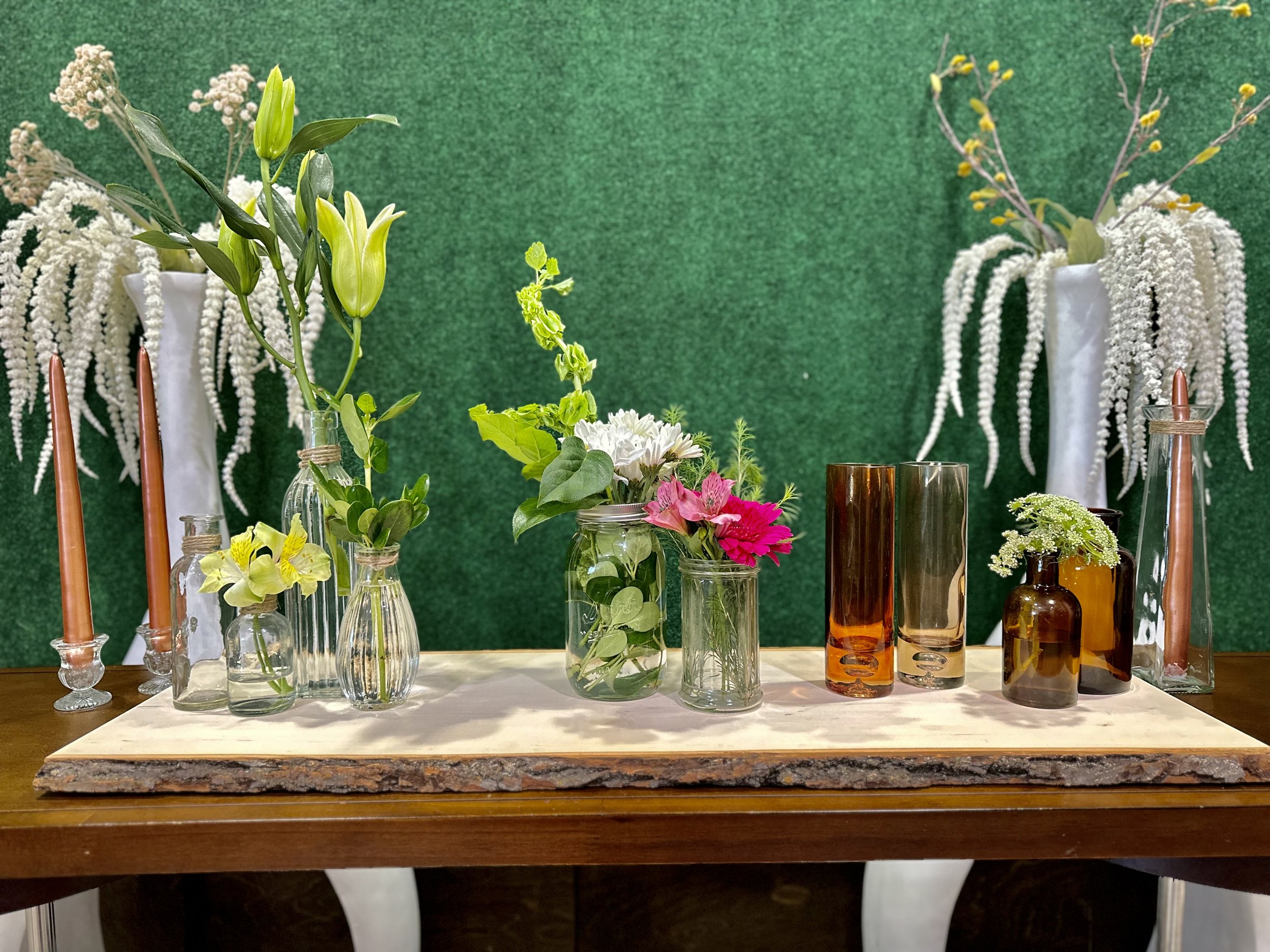 A table decorated with various glass vases containing different types of flowers, including lilies, daisies, and greenery. The backdrop is a green textured surface, and the vases are surrounded by two long orange candles and white floral arrangements in tall white vases.