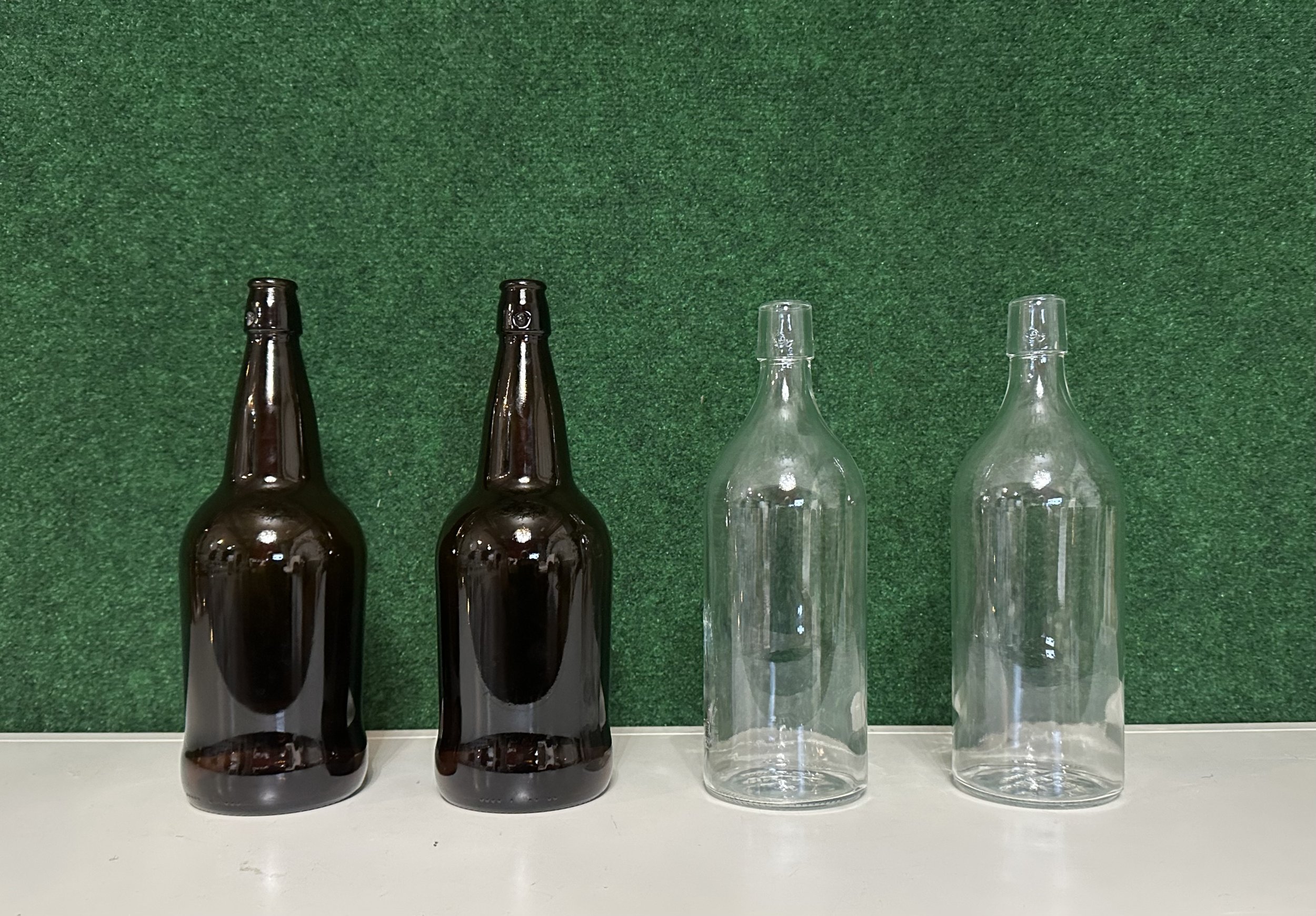 Four empty glass bottles on a white surface, two brown and two clear, against a green background.