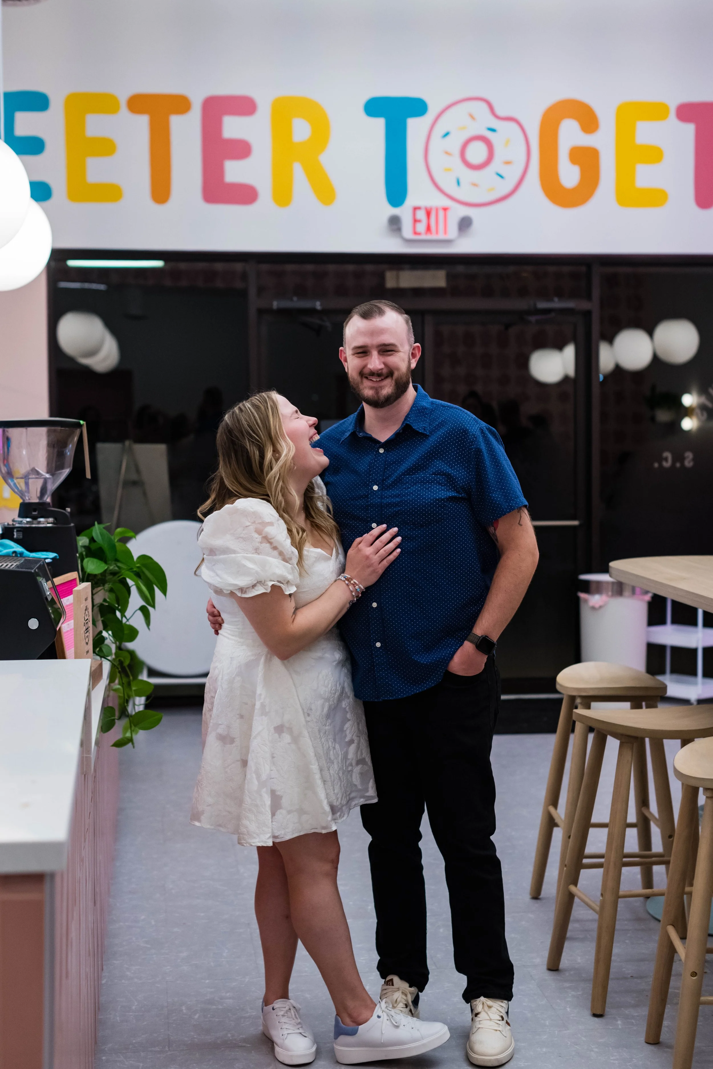 A woman and a man standing inside an ice cream shop, smiling and enjoying each other's company. The shop has colorful signage with the word 'TOGETHER' and a donut graphic in the background.