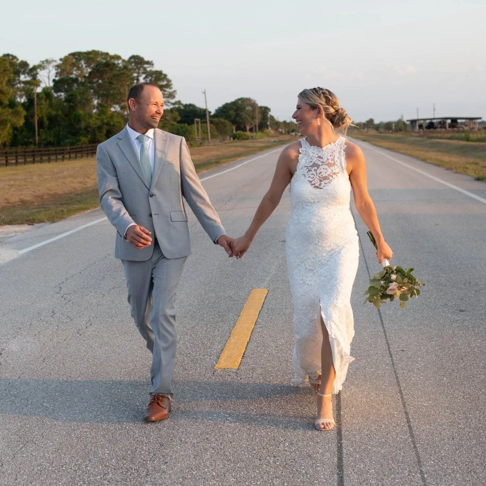 A bride and groom walking hand in hand on an empty rural road during sunset, smiling and looking at each other.