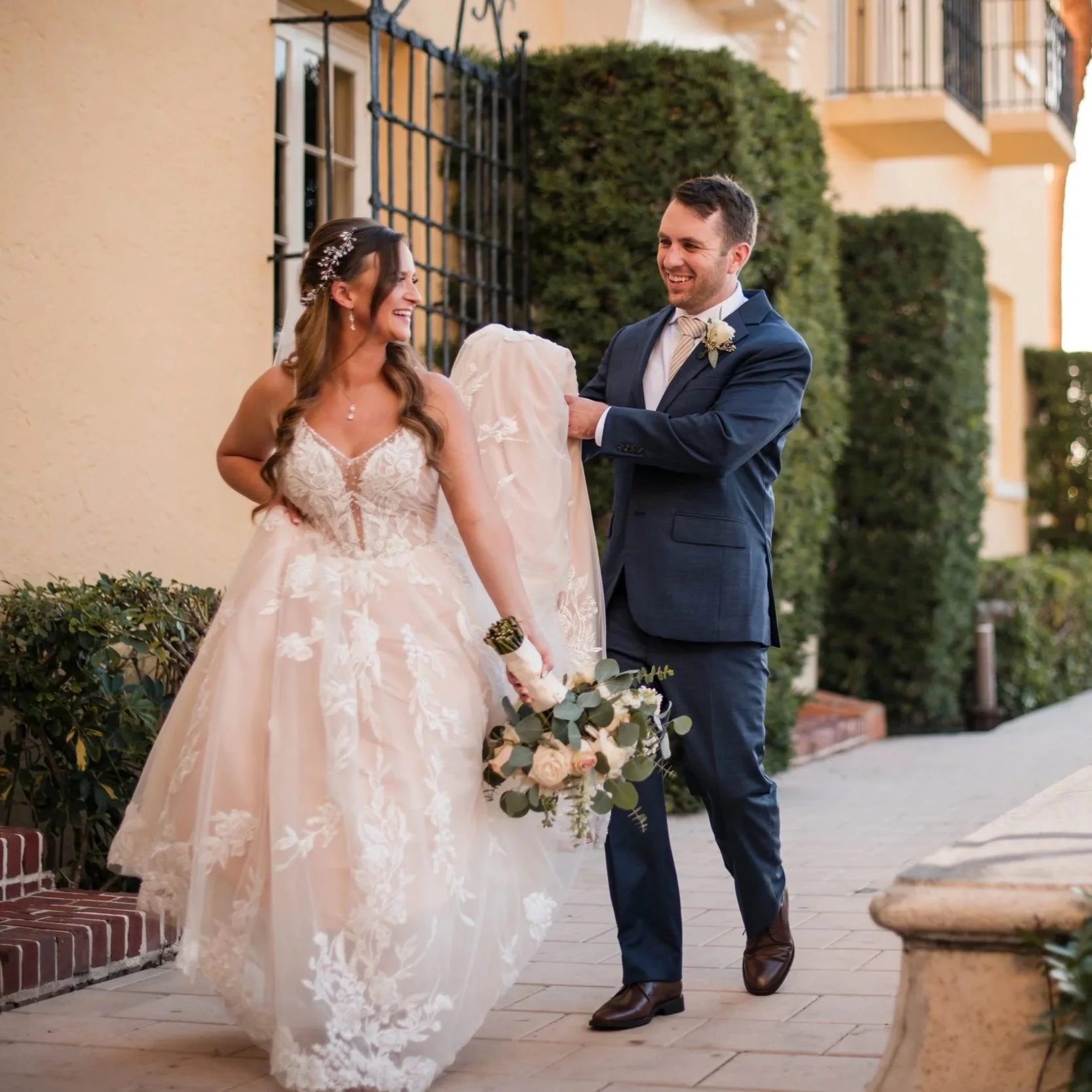 A bride in a white lace wedding gown holding a bouquet of flowers and a groom in a navy suit smiling at each other as the groom adjusts the bride's dress outside a building with greenery.