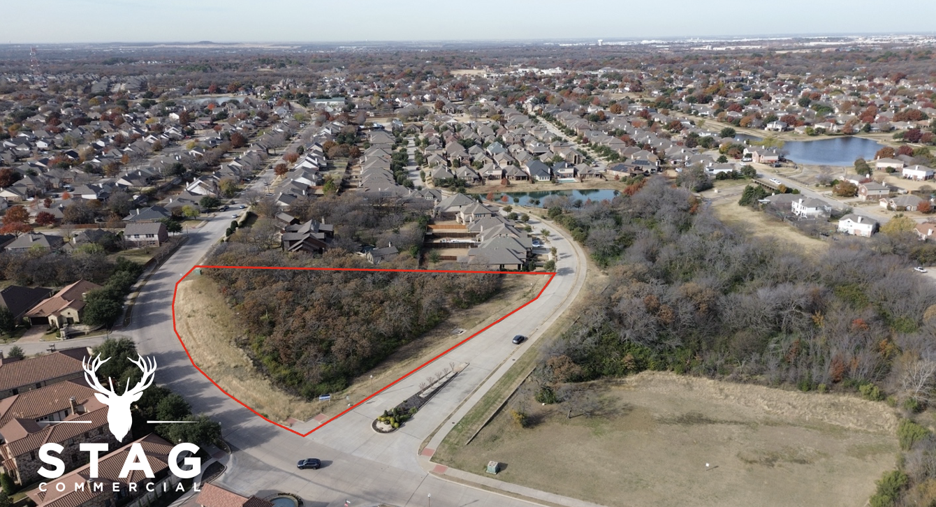 An aerial view of a residential neighborhood with a cleared lot outlined in red, showing a mix of trees and grass, with houses, roads, and a small pond in the background.