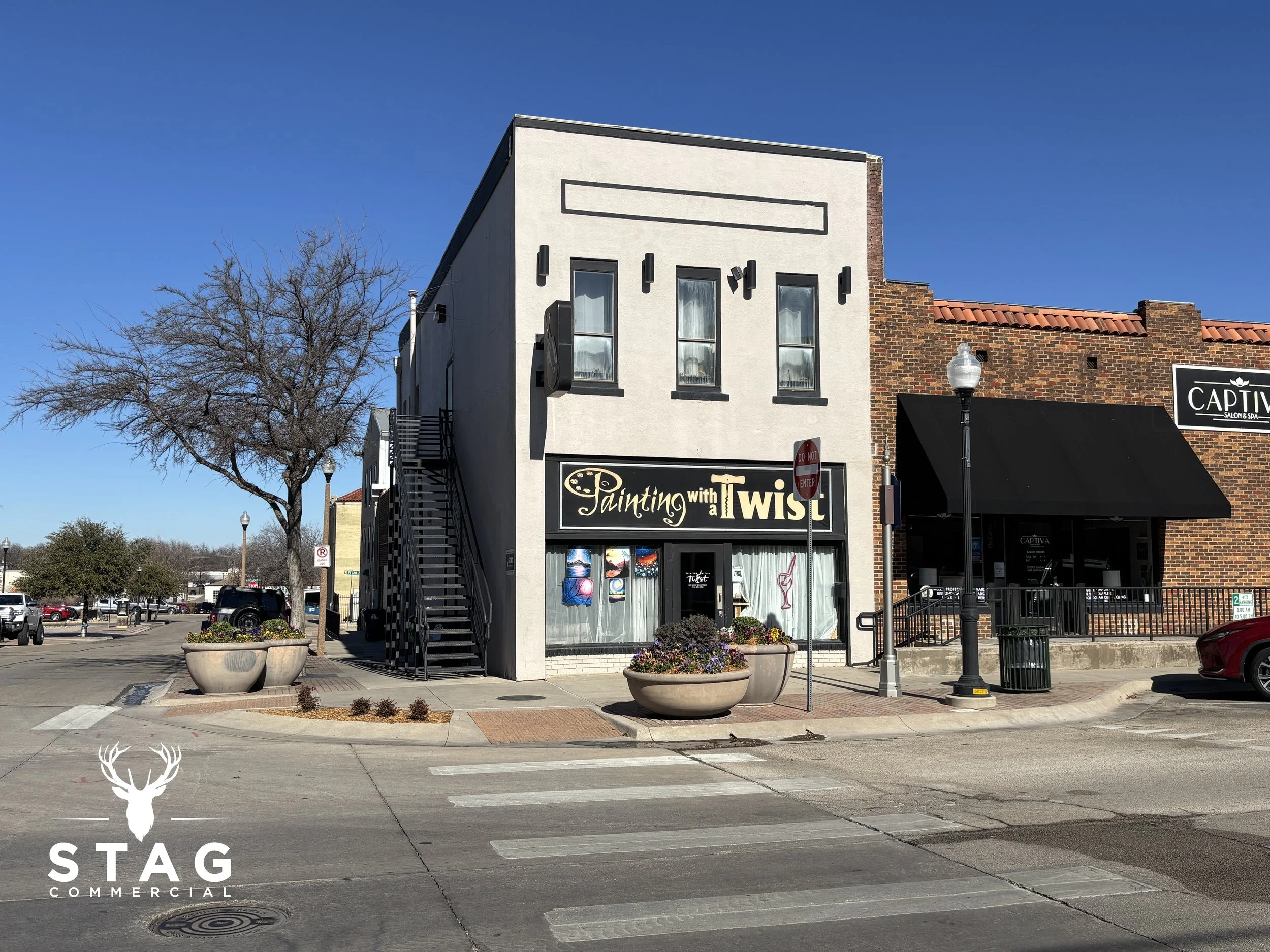 A two-story commercial building on a city street, with a sign reading "Painting with a Twist" on the front, large windows with art displays, and an exterior staircase on the left side. There are flower pots on the sidewalk and a tree nearby, with parking and other storefronts in the background under a clear blue sky.
