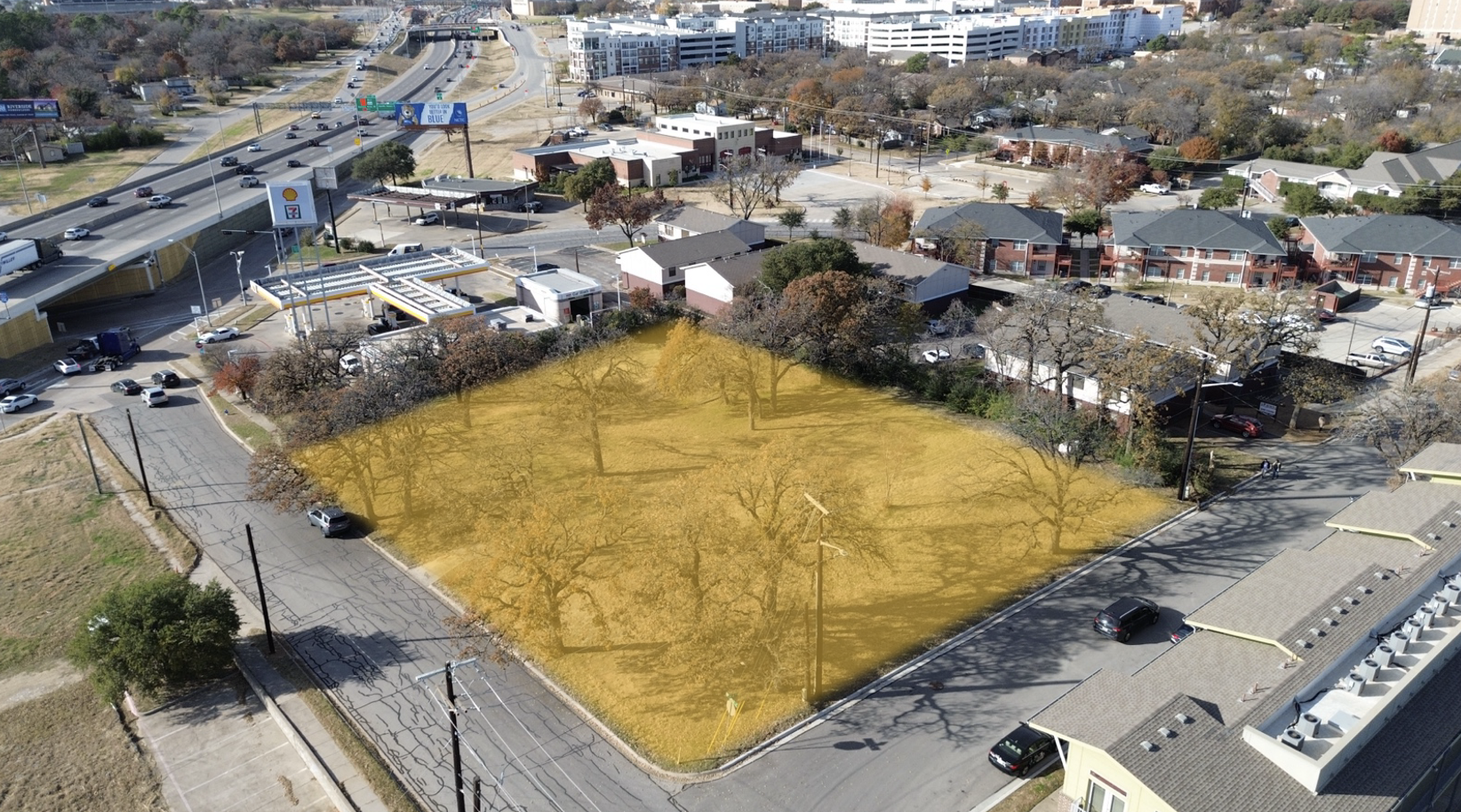 An aerial view of a cityscape featuring a highlighted empty lot with a few trees, surrounded by residential and commercial buildings, roads, and parking lots.
