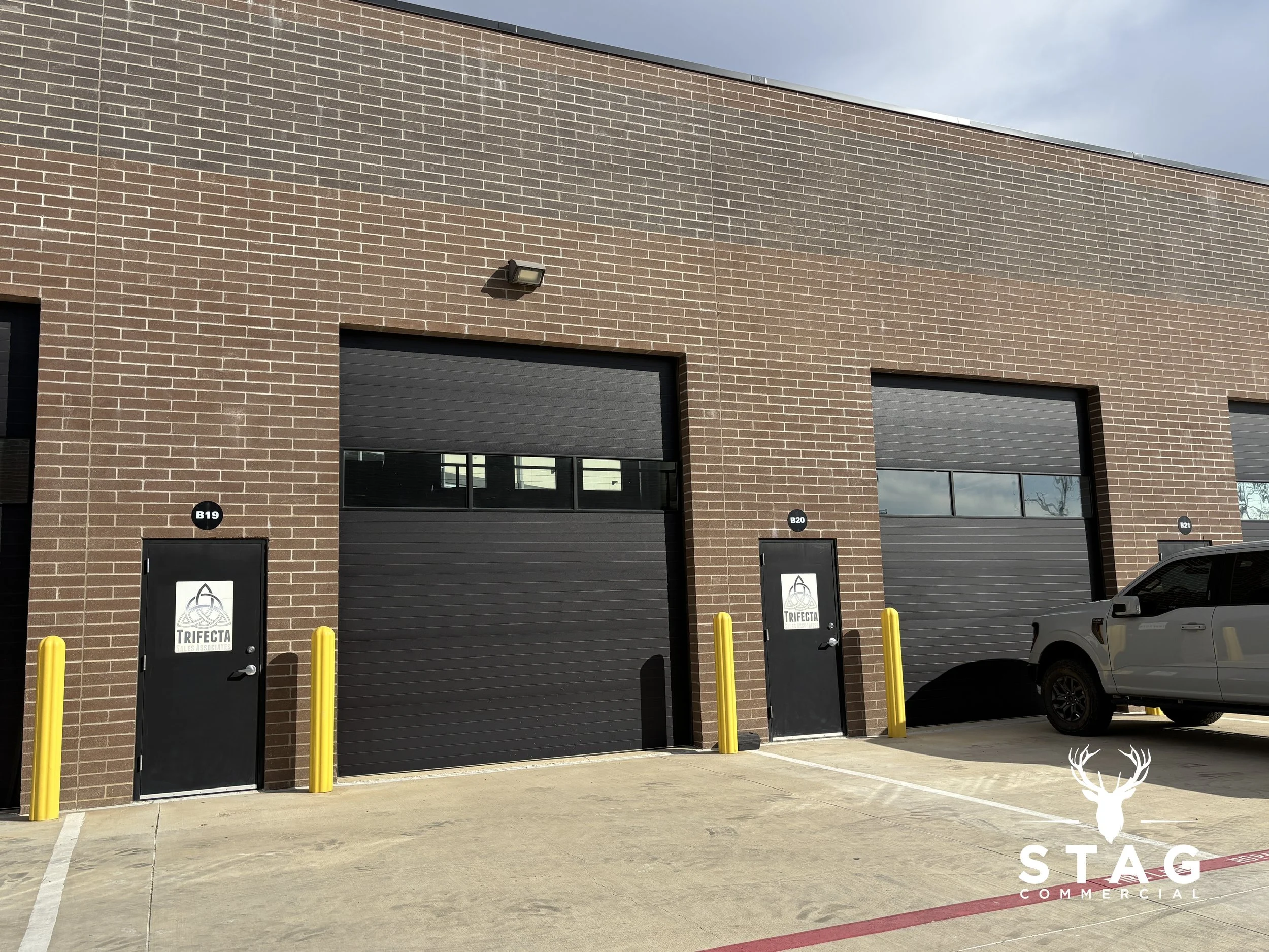 Commercial building with brick exterior, featuring two black garage doors with windows, two black doors with 'TRIFECTA' logos, and a parked gray truck in front. Yellow safety bollards are placed near the doors.