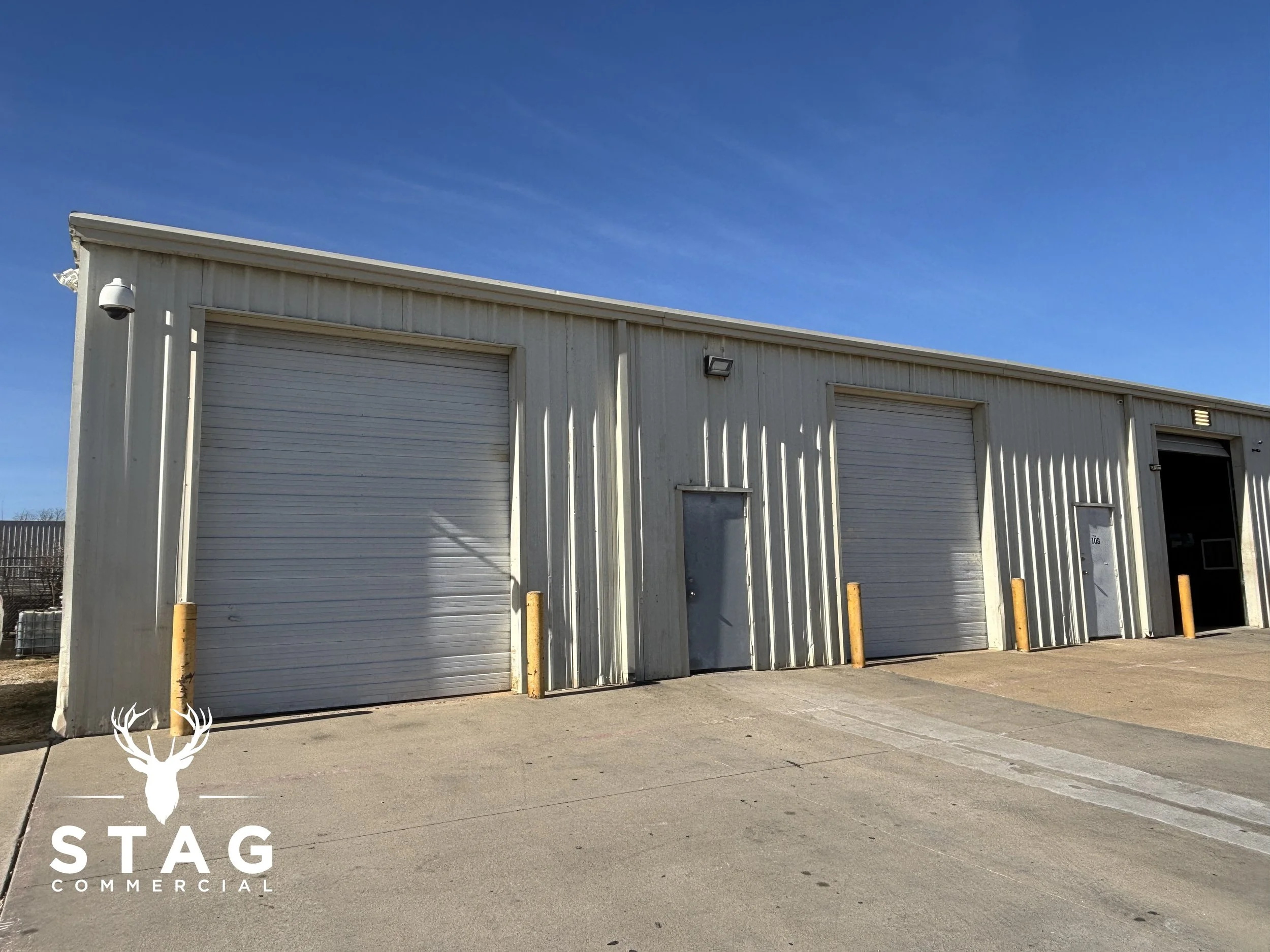 Exterior view of a beige industrial warehouse with three large roll-up garage doors under a clear blue sky.