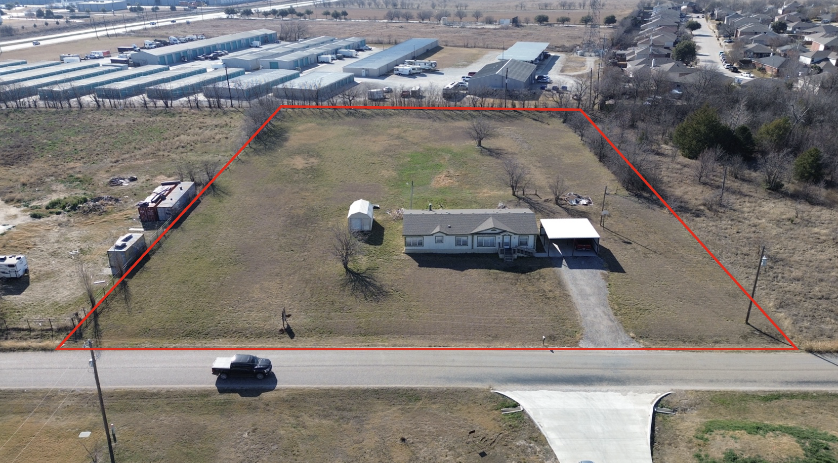 Aerial view of a large lot with a house, a small shed, and a carport, outlined in red, next to a road with a black truck. Surrounding areas include a row of commercial buildings and residential neighborhood.