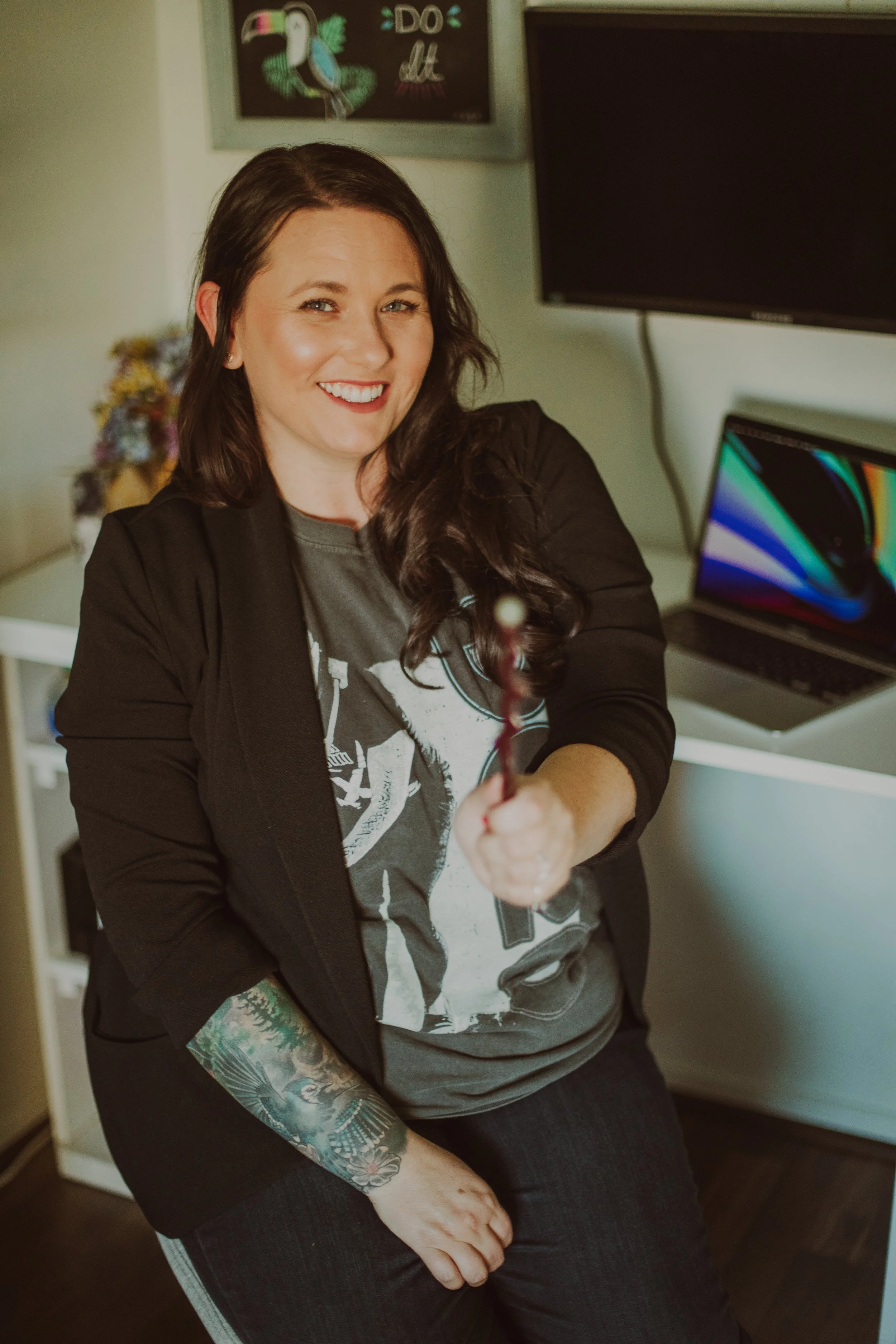 Young female therapist in a band tee sitting in front of a desk with laptop open waving a magic wand.