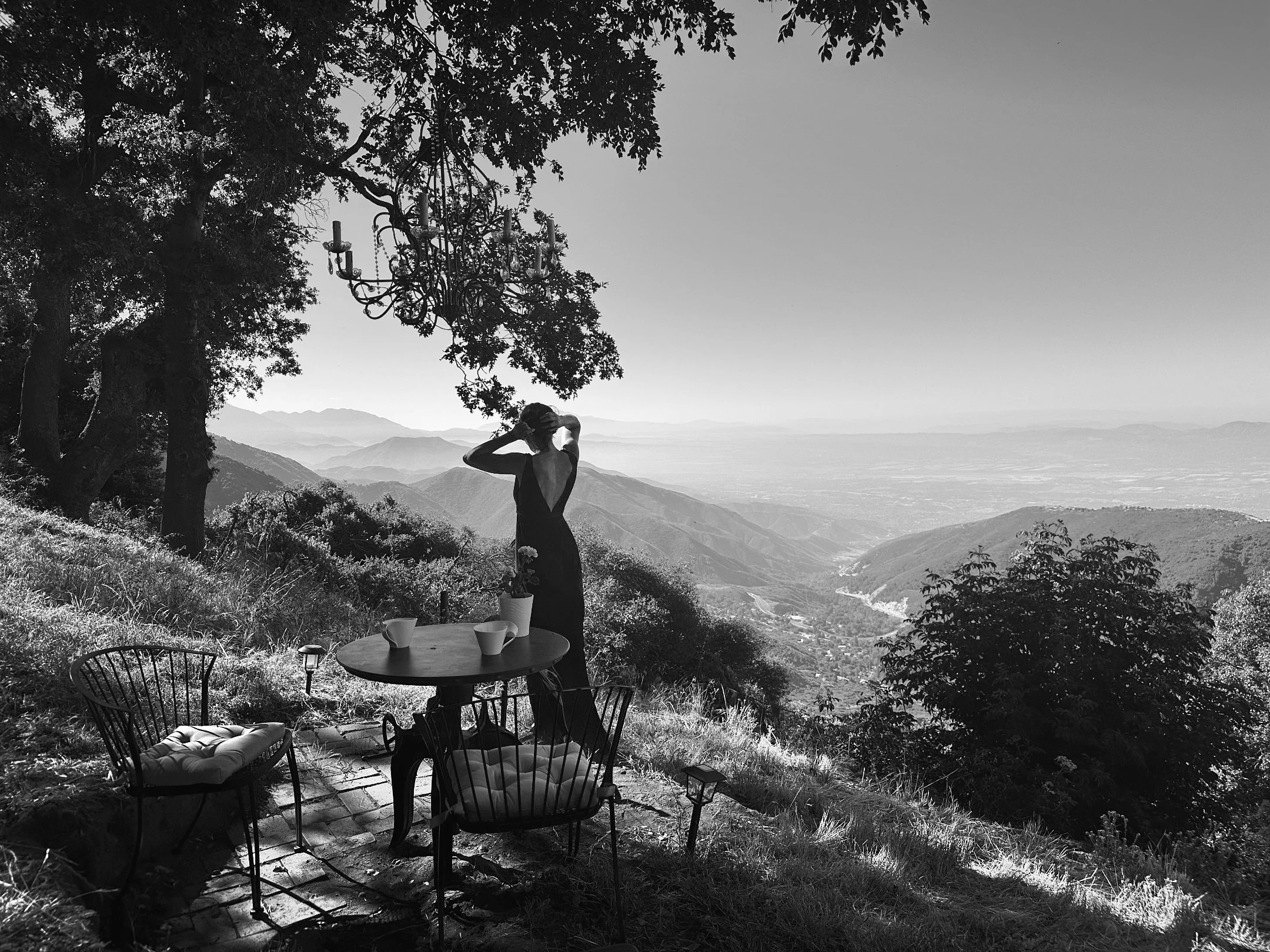 Host Anita Hodson at Holly Hill Chalet in Crestline, California—captured in black and white in the garden overlooking the San Bernardino Mountains, with a chandelier hanging from an oak tree and cinematic light that makes the chalet a photographer’s 